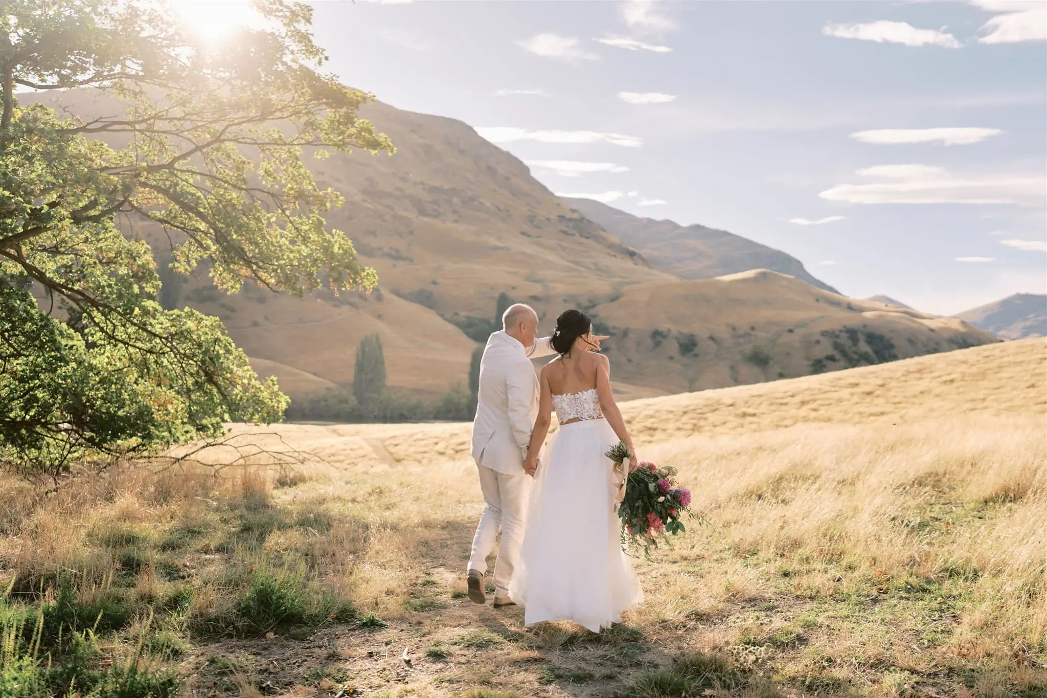 queenstown new zealand heli wedding elopement photographer videographer | A bride and groom walk hand in hand through a sunlit field, the backdrop of rolling hills setting the perfect scene for their Queenstown elopement, the bride carrying a bouquet.