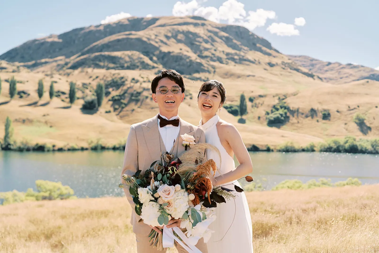 queenstown new zealand heli wedding elopement photographer videographer | A newlywed couple smiling joyfully, holding a bouquet with a scenic lake and sunlit hills in the background during their Queenstown Pre-Wedding Shoot.