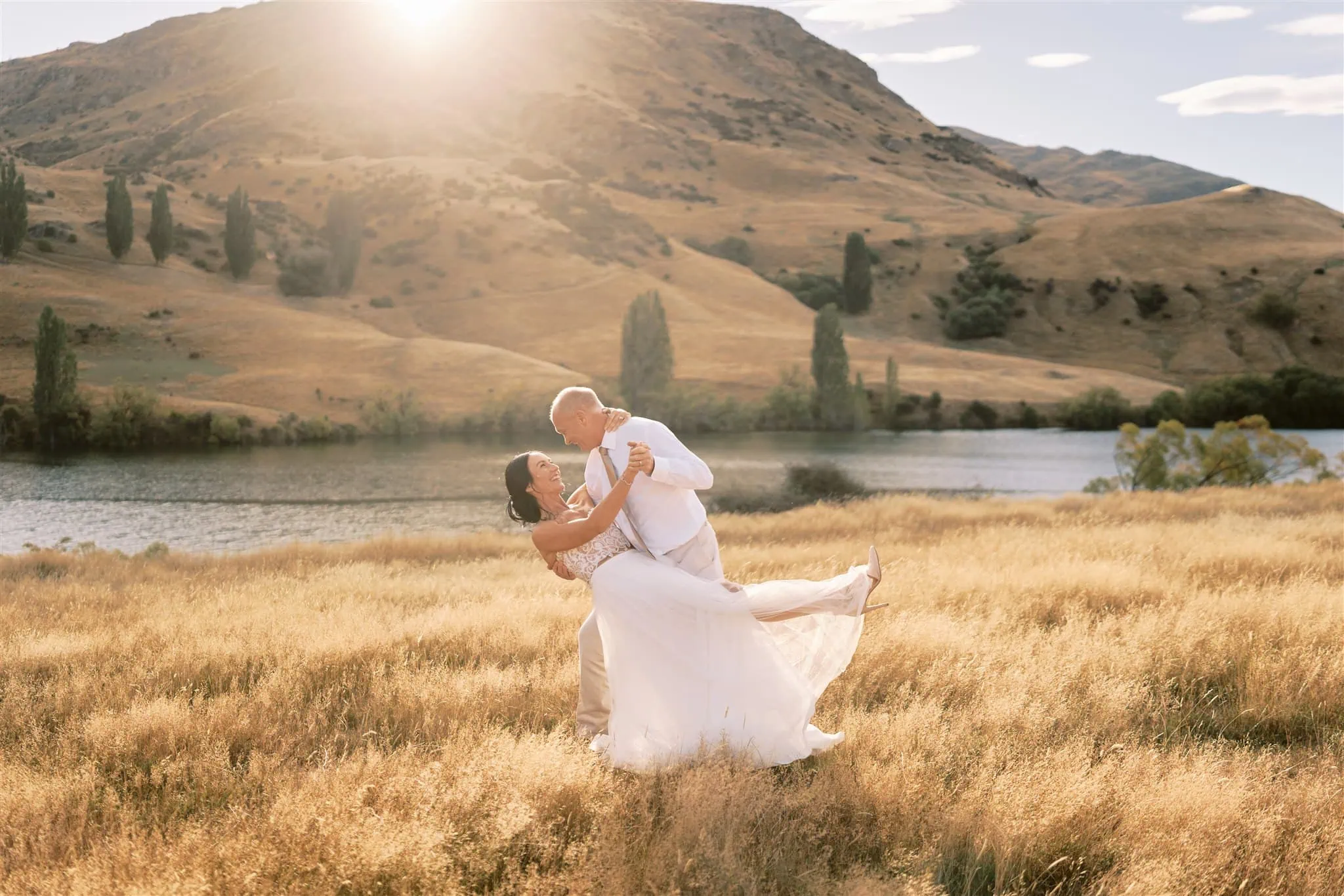 queenstown new zealand heli wedding elopement photographer videographer | Ian & Vivienne, a couple in wedding attire, dancing in a sunlit field near a lake with rolling hills in the background.