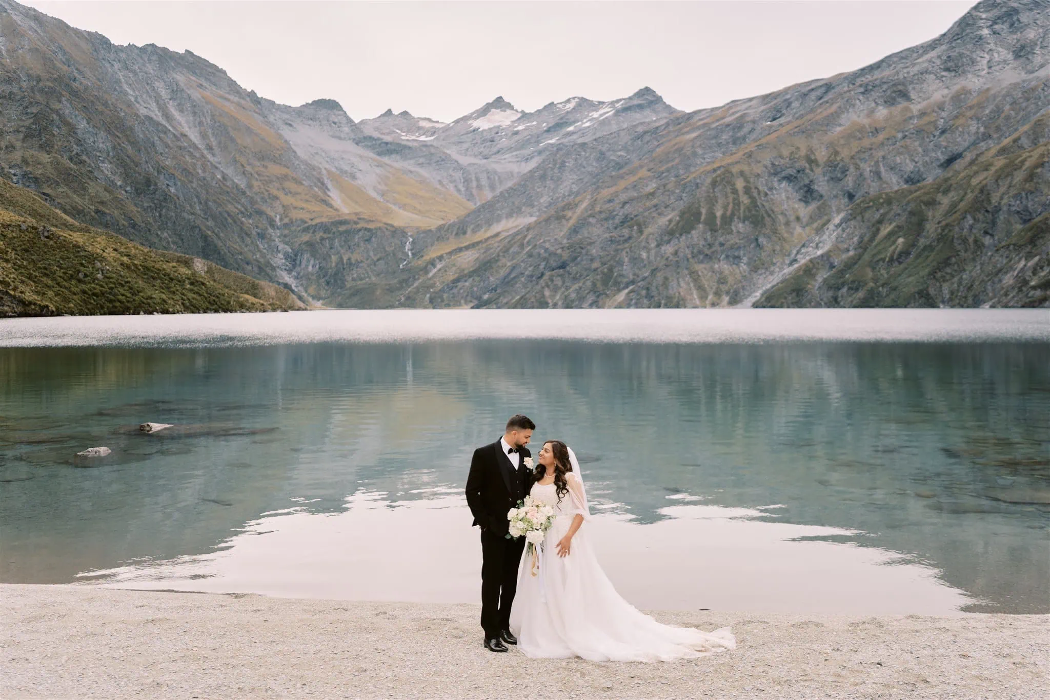 queenstown new zealand heli wedding elopement photographer videographer | VidhyA and Kartik embrace on a lakeshore with a mountain backdrop during their Queenstown elopement.