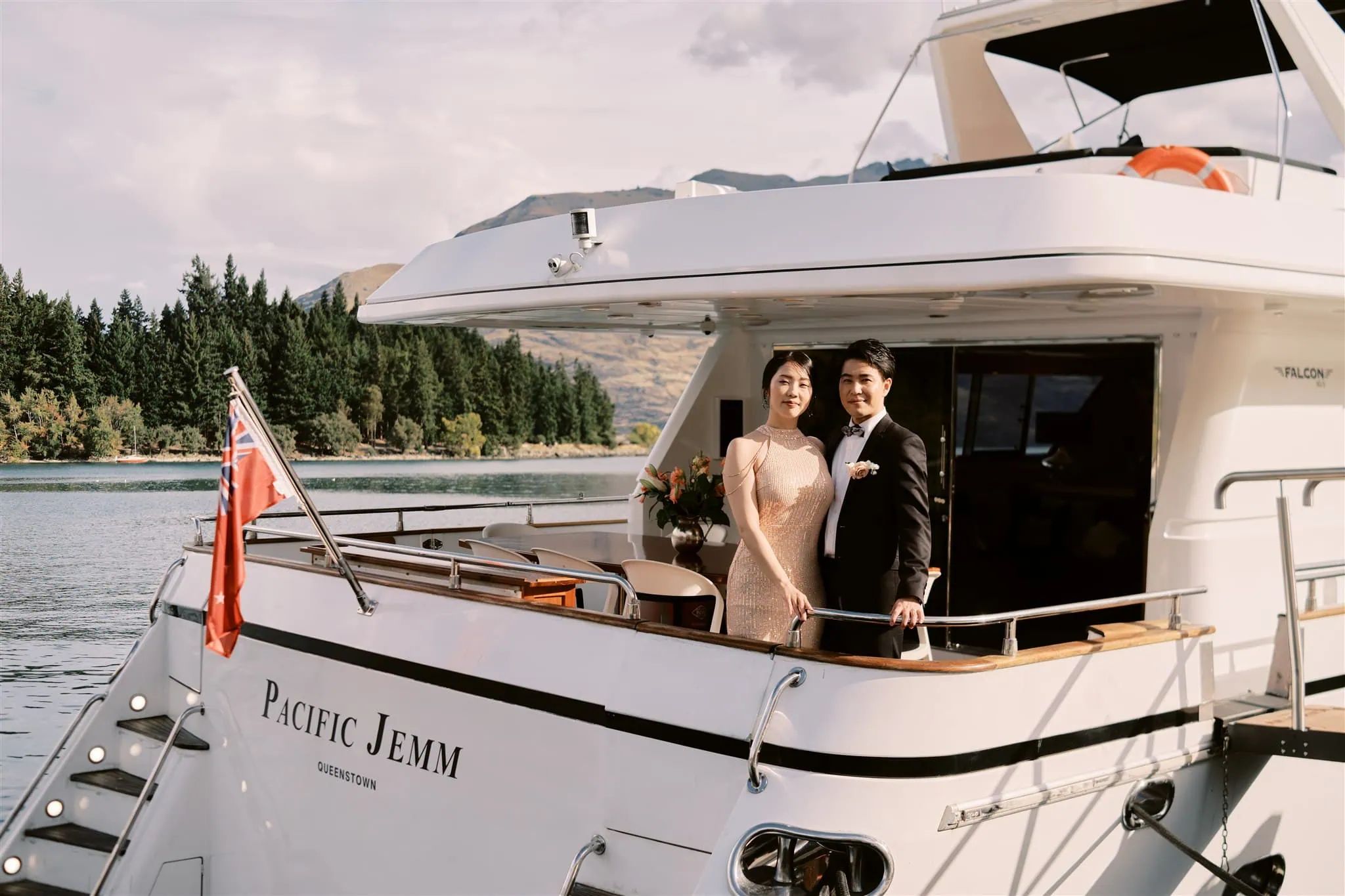 queenstown new zealand heli wedding elopement photographer videographer | A couple in formal wedding attire posing on the deck of the yacht "Pacific Jemm" with a scenic lake and mountains in the background during their Queenstown Elopement.