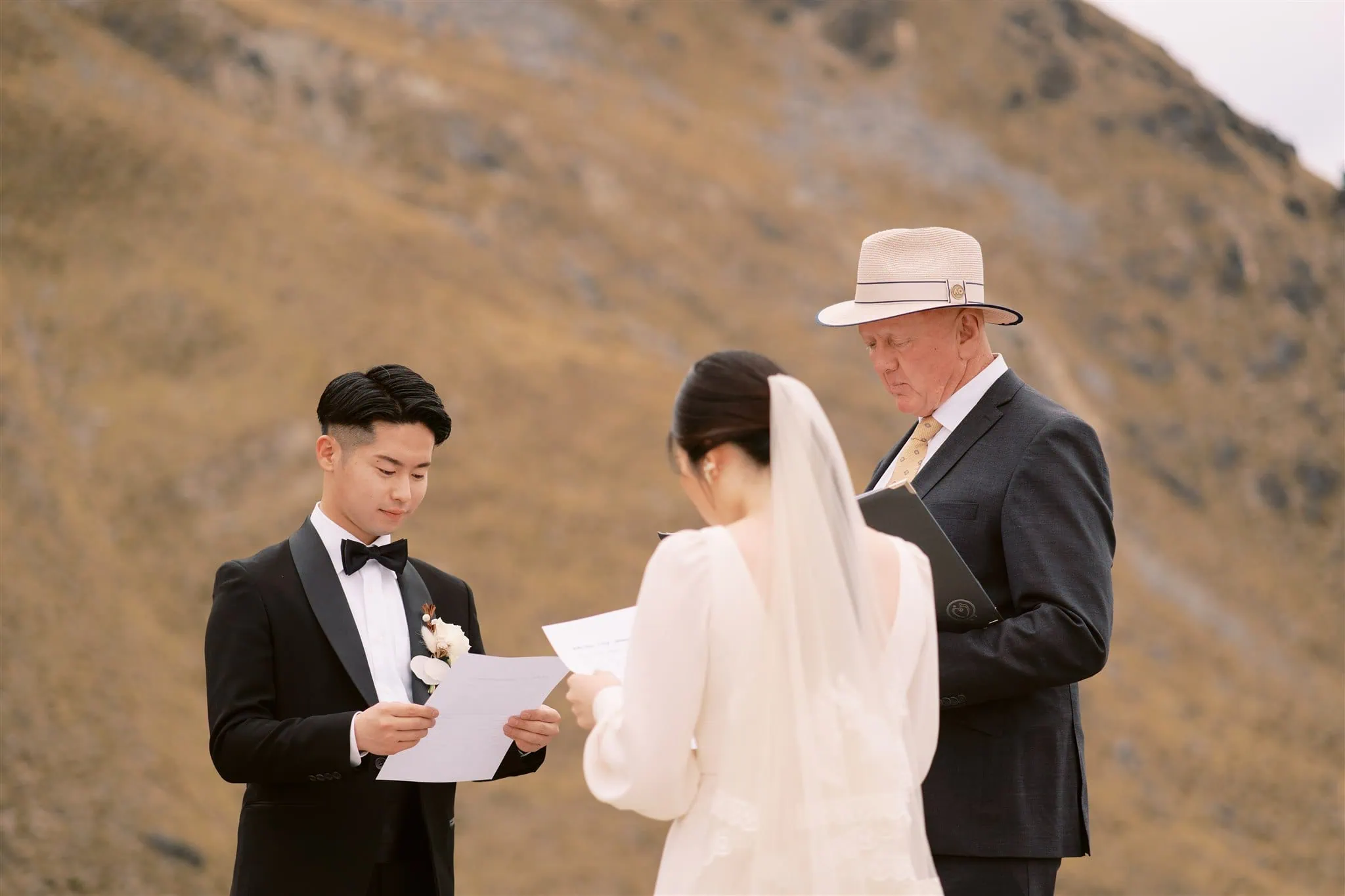 queenstown new zealand heli wedding elopement photographer videographer | A couple exchanges vows during an outdoor wedding ceremony officiated by a man in a hat, with a mountainous landscape in the background, all beautifully captured by a Queenstown wedding photographer.