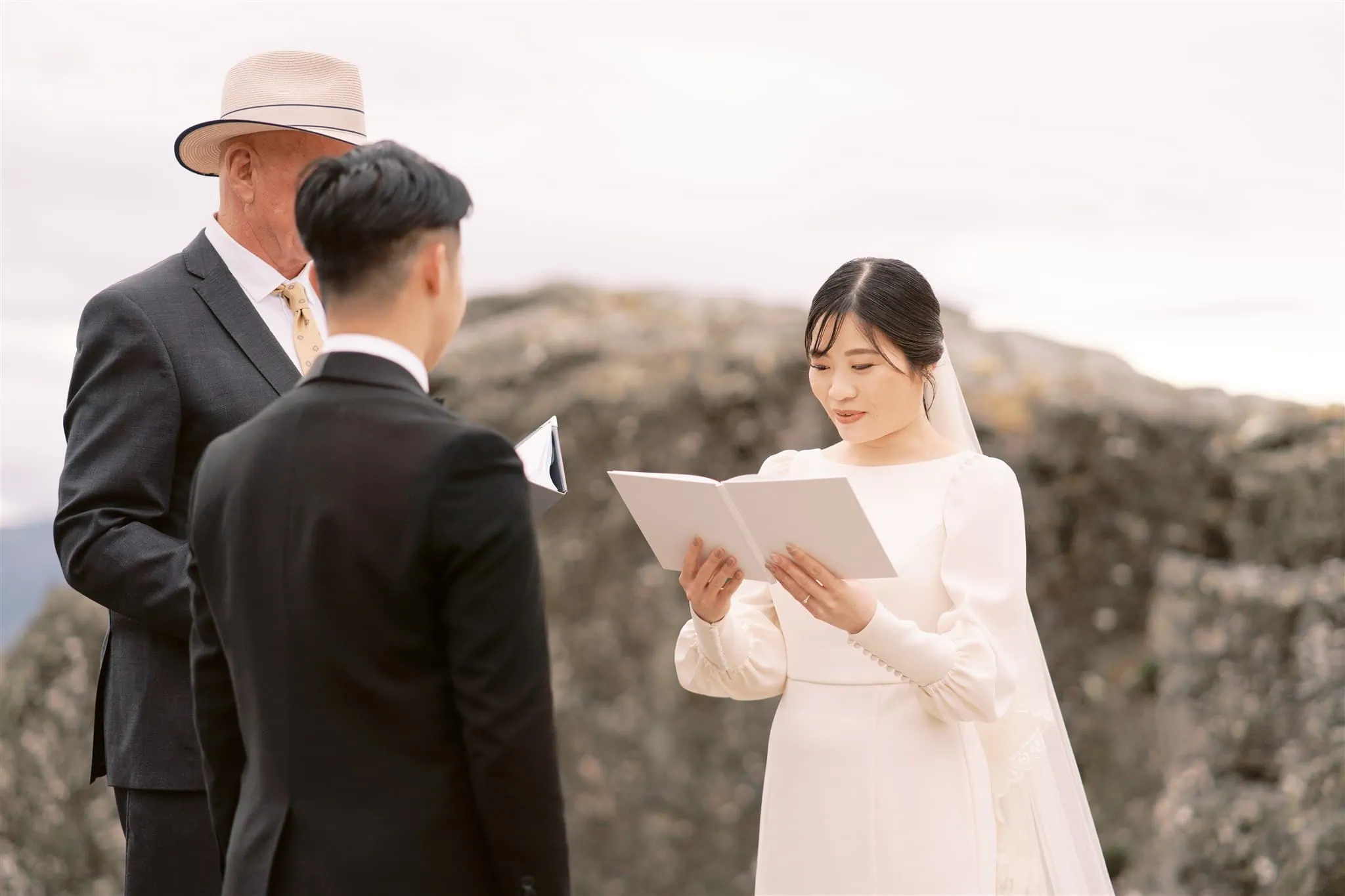 queenstown new zealand heli wedding elopement photographer videographer | A bride in a white dress reads from a book during an outdoor wedding ceremony, captured beautifully by a Queenstown wedding photographer. The groom in a black suit and an officiant in a suit and hat stand nearby, with rocks and a cloudy sky forming the picturesque backdrop.