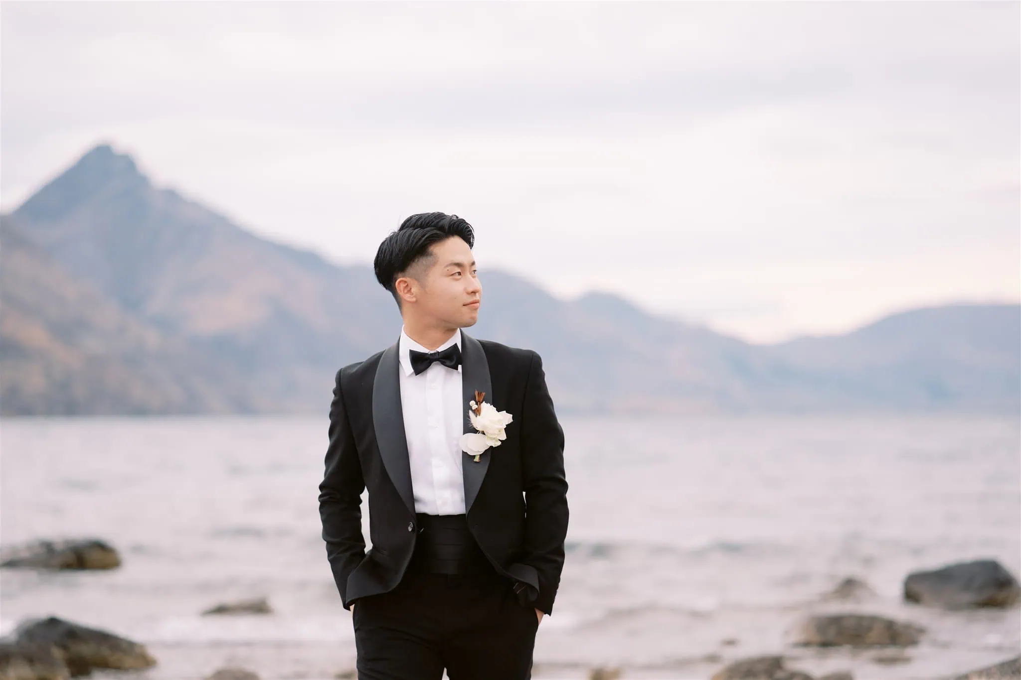 queenstown new zealand heli wedding elopement photographer videographer | A man in a black tuxedo with a white boutonniere stands near a rocky shoreline with mountains in the background, perfectly captured by a Queenstown wedding photographer.