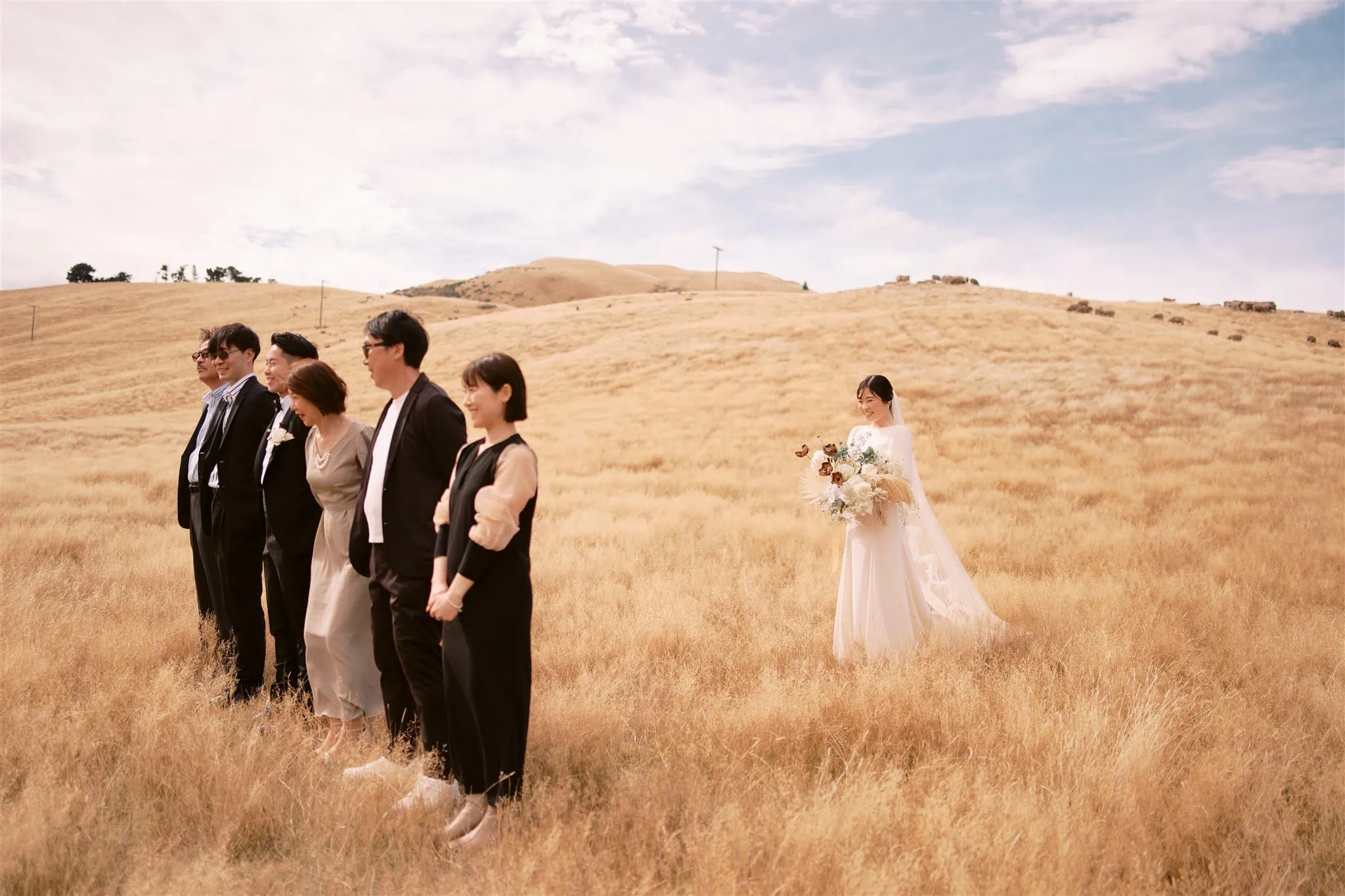 queenstown new zealand heli wedding elopement photographer videographer | A bride in a white dress holding a bouquet stands in a golden field; five people dressed in formal attire stand in a line nearby, facing sideways, under a partly cloudy sky. Captured perfectly by the Queenstown wedding photographer.