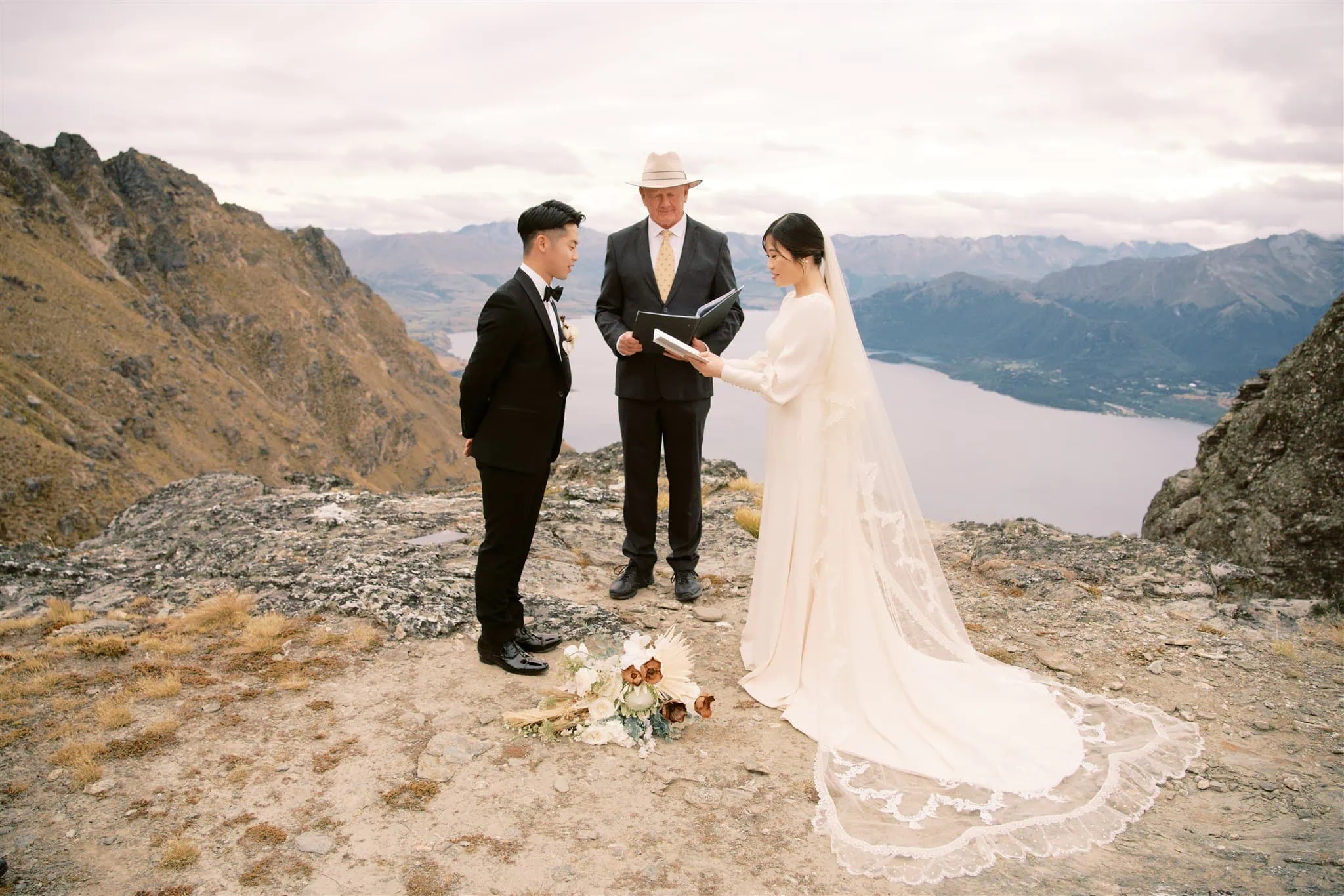 queenstown new zealand heli wedding elopement photographer videographer | A couple in wedding attire stands on a rocky mountain ledge, exchanging vows with an officiant. The backdrop includes a lake and mountain range under an overcast sky. Captured by a Queenstown wedding photographer, a floral arrangement lies gracefully on the ground.