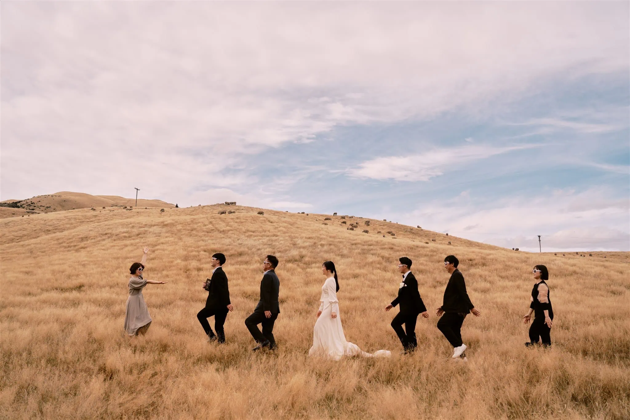 queenstown new zealand heli wedding elopement photographer videographer | A wedding party walks through a grassy field with hills in the background, captured beautifully by a Queenstown wedding photographer. The bride and groom are at the center, accompanied by their attendants dressed in formal attire.