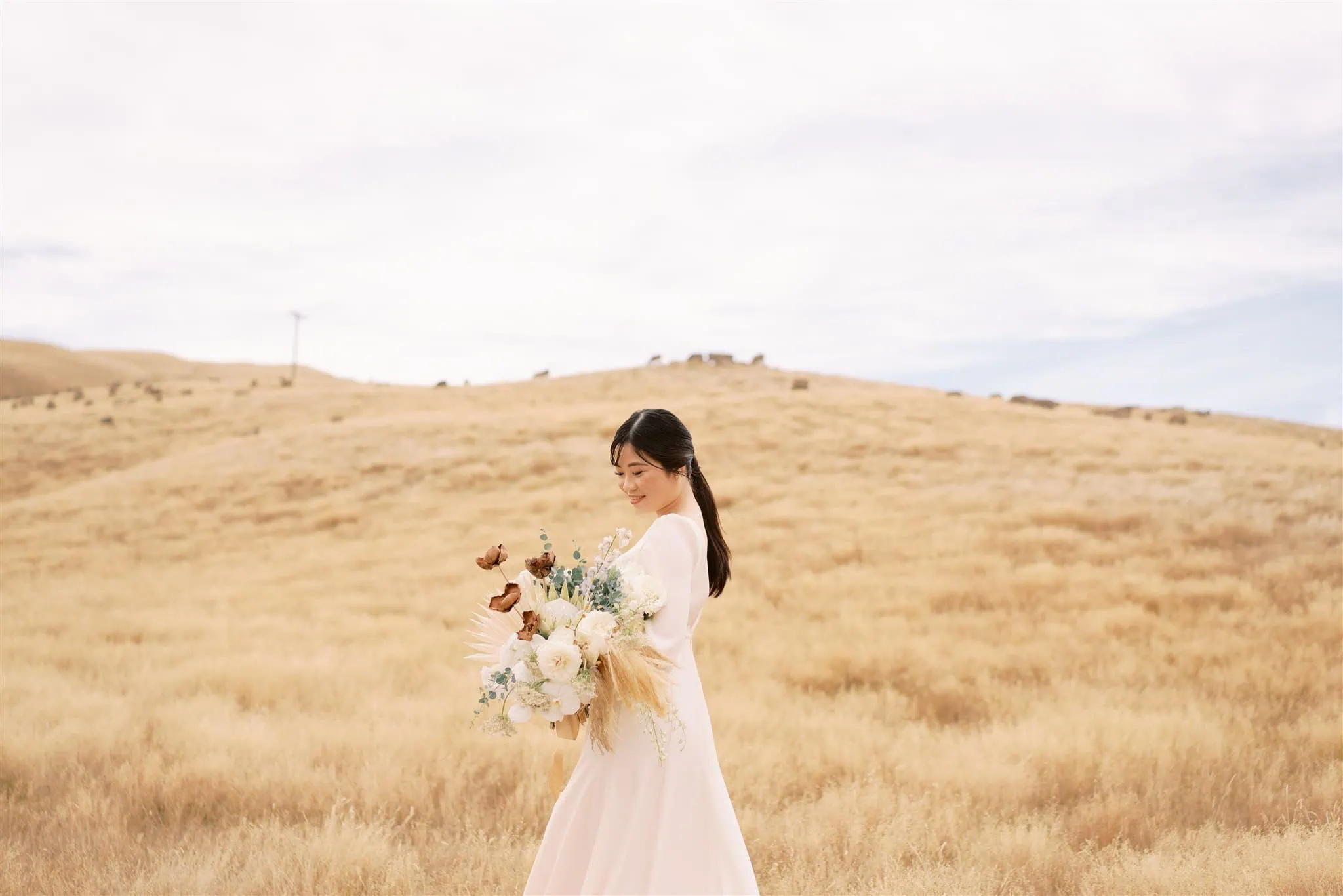 queenstown new zealand heli wedding elopement photographer videographer | A woman in a light-colored dress holds a bouquet of flowers while standing in an open field with dry grass under a cloudy sky, captured beautifully by Queenstown wedding photographer.