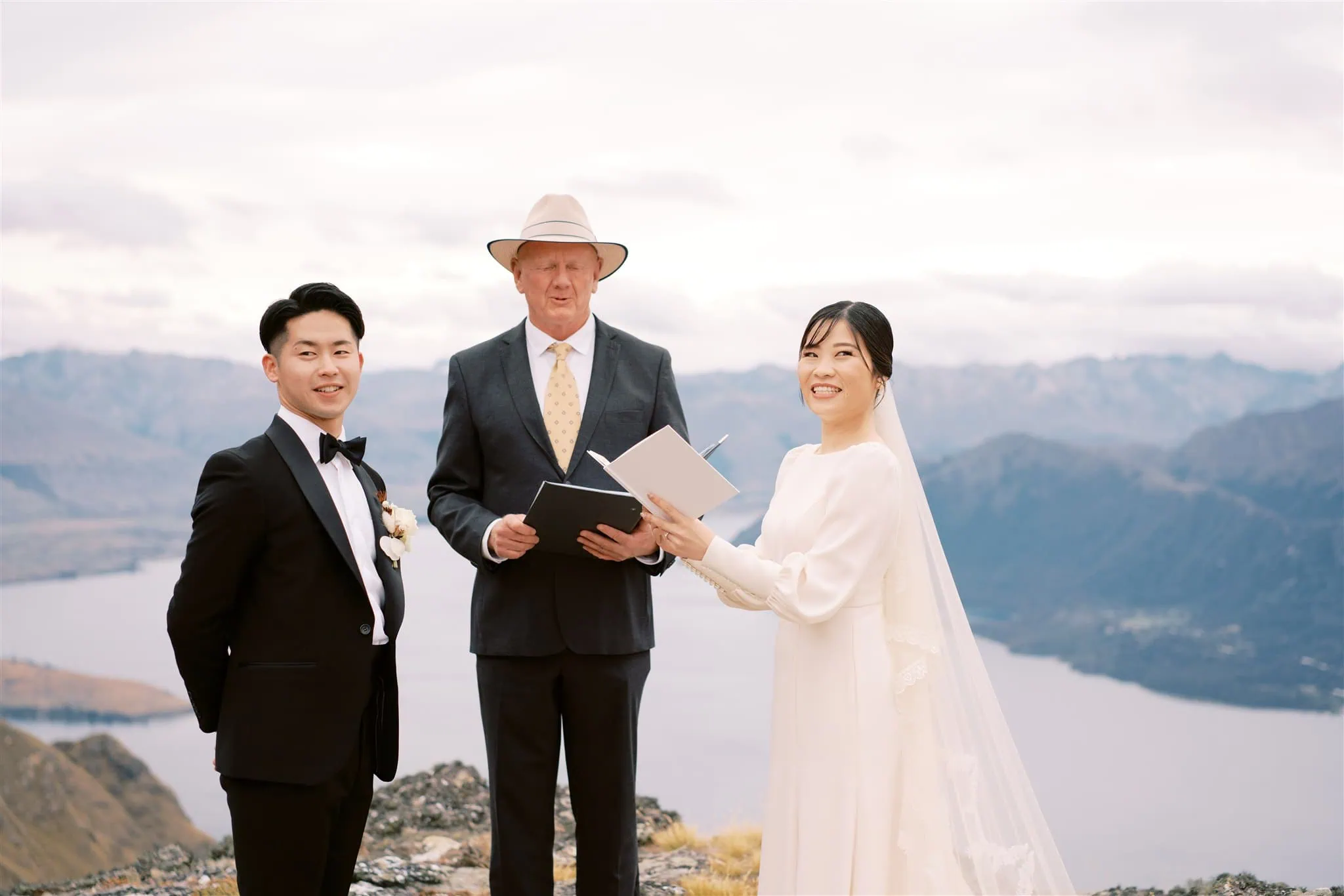 queenstown new zealand heli wedding elopement photographer videographer | A bride and groom stand with an officiant on a mountain overlook during their wedding ceremony, captured beautifully by a Queenstown wedding photographer. The bride holds a booklet and smiles, while the groom stands with hands in his pockets.