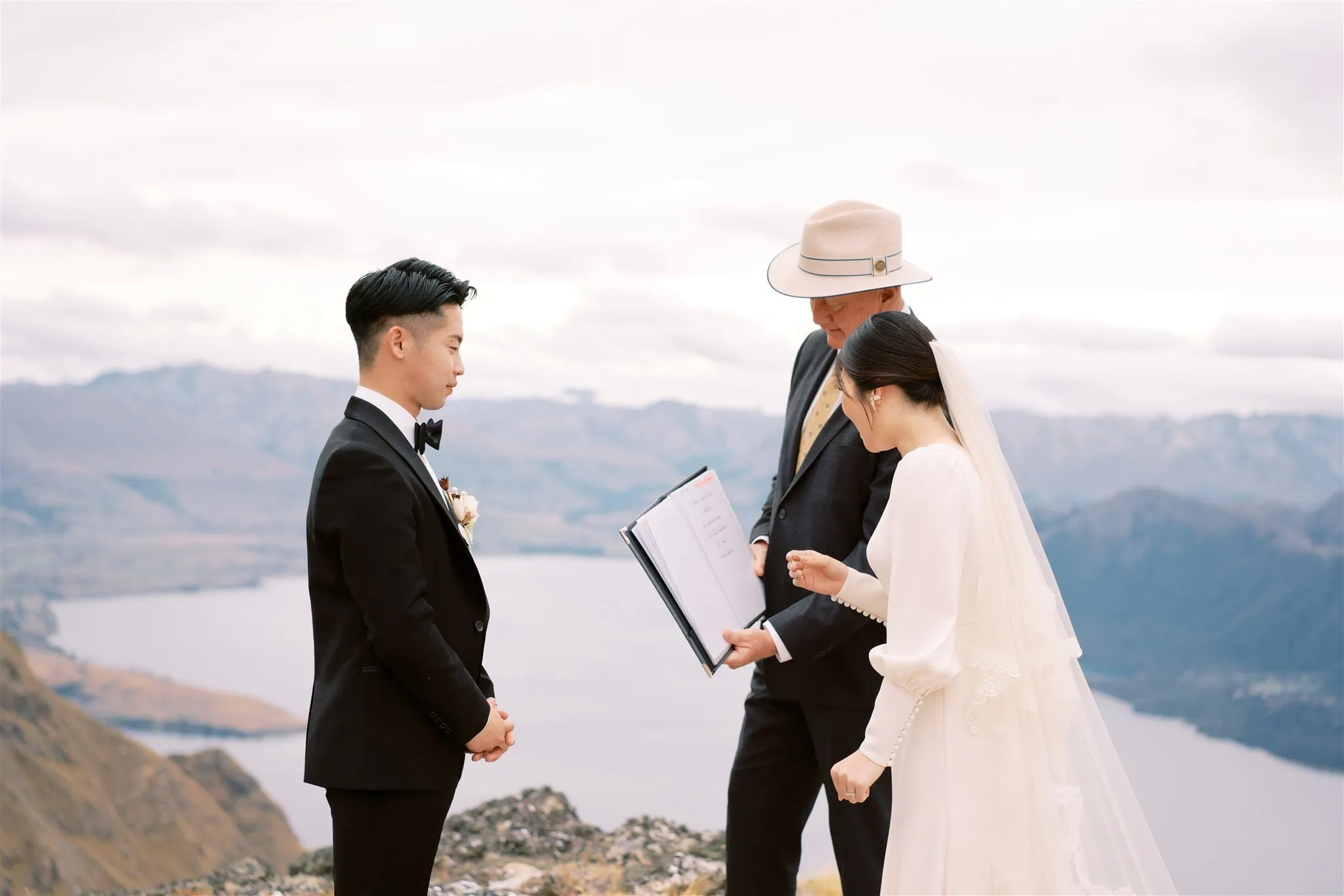 queenstown new zealand heli wedding elopement photographer videographer | A couple stands with an officiant during a wedding ceremony on a mountain top with a scenic view of a lake and mountains in the background, captured beautifully by their Queenstown wedding photographer.