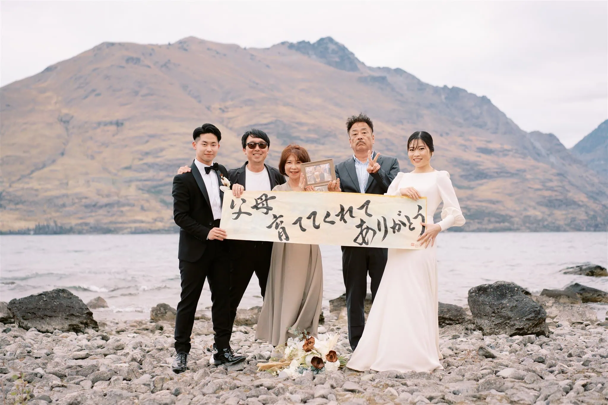 queenstown new zealand heli wedding elopement photographer videographer | A group of five people in formal attire stands on a rocky shoreline holding a large banner with Japanese text. A mountain and lake are in the background. One person, possibly the Queenstown wedding photographer, is holding a framed photo to capture this unique moment.