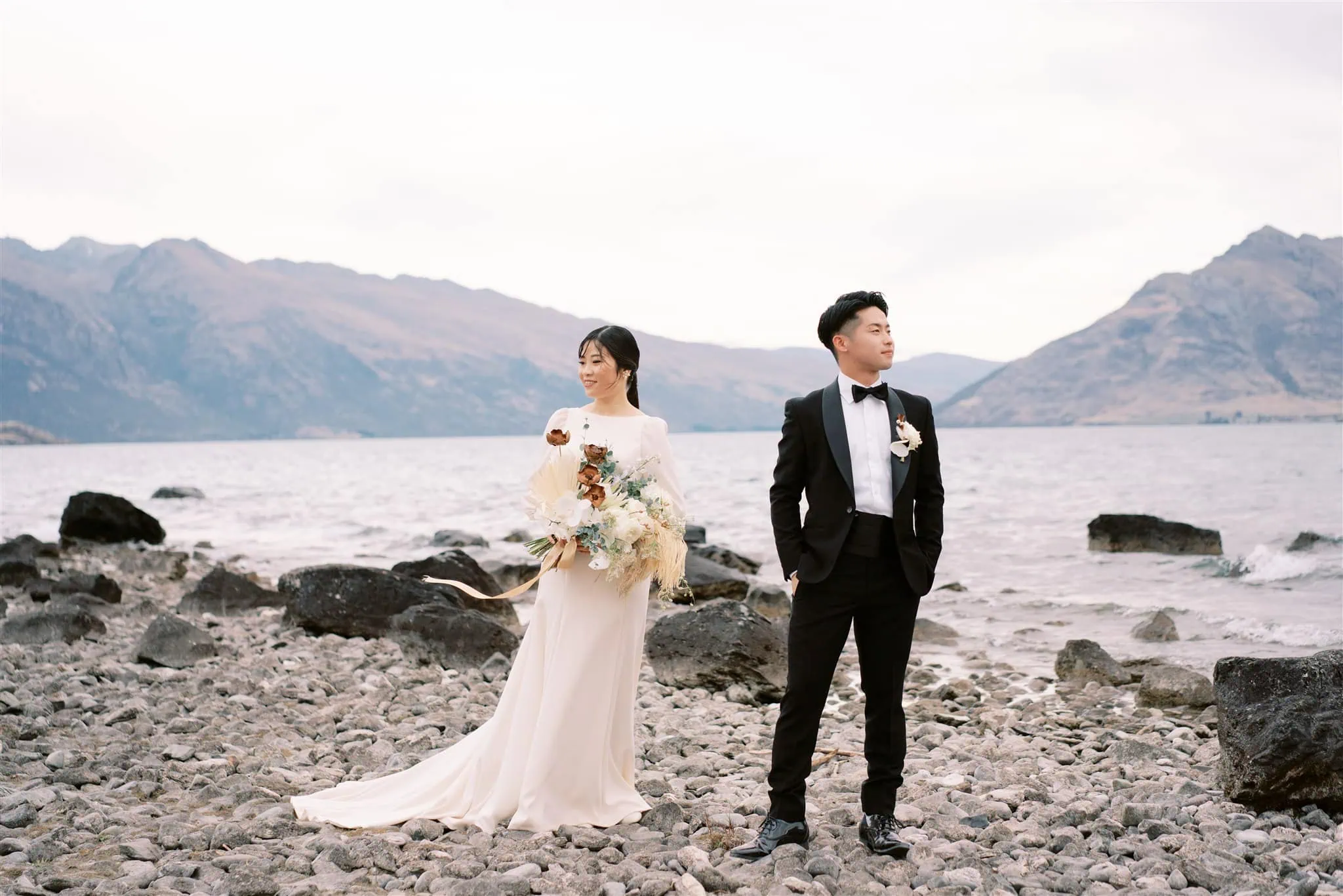 queenstown new zealand heli wedding elopement photographer videographer | A bride in a white dress holds a bouquet and stands beside a groom in a black suit on a rocky lakeshore with mountains in the background, captured beautifully by their Queenstown wedding photographer.
