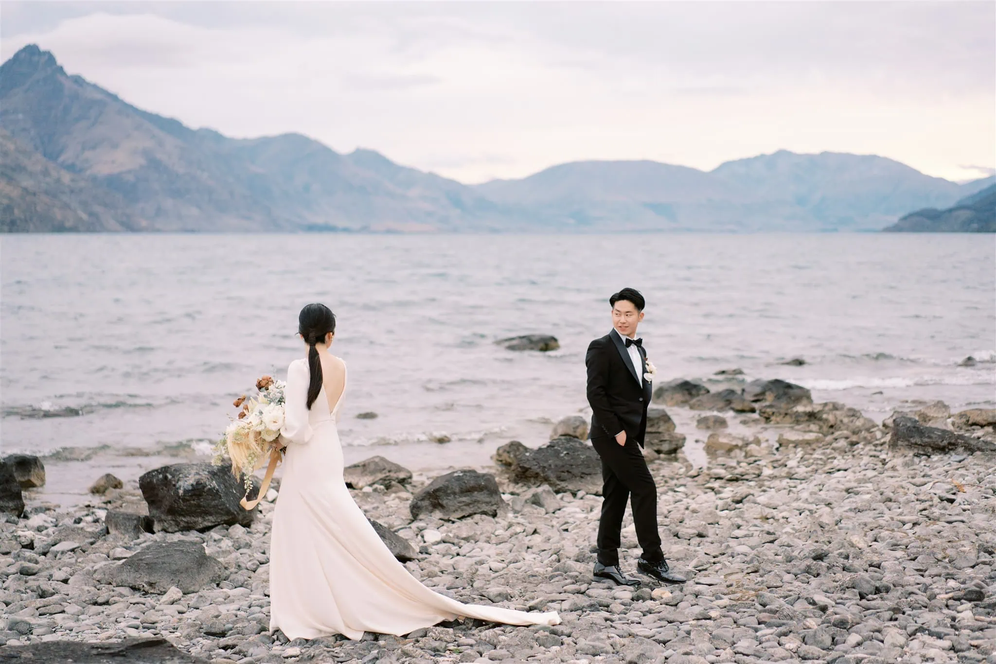 queenstown new zealand heli wedding elopement photographer videographer | A couple dressed in wedding attire stands on a rocky shore by a lake, with majestic mountains in the background. The bride holds a bouquet and gazes lovingly at the groom. Captured perfectly by a Queenstown wedding photographer, this moment is as timeless as the scenery around them.