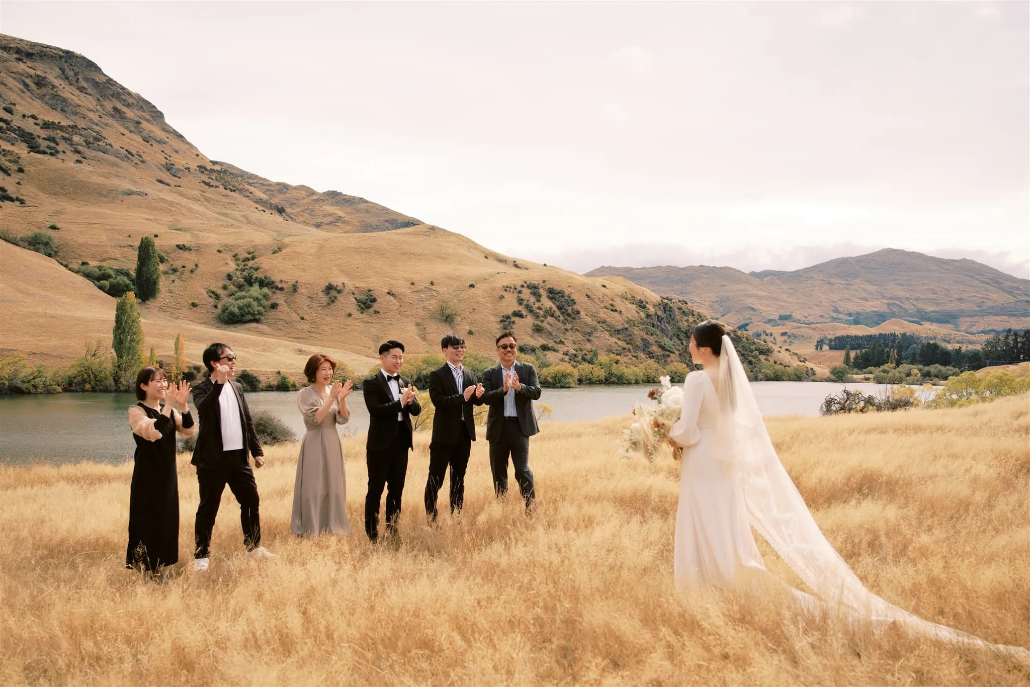 queenstown new zealand heli wedding elopement photographer videographer | A Queenstown wedding photographer captures a bride in a white dress, standing before five people who are clapping and smiling, with grassy hills and a serene river in the background.