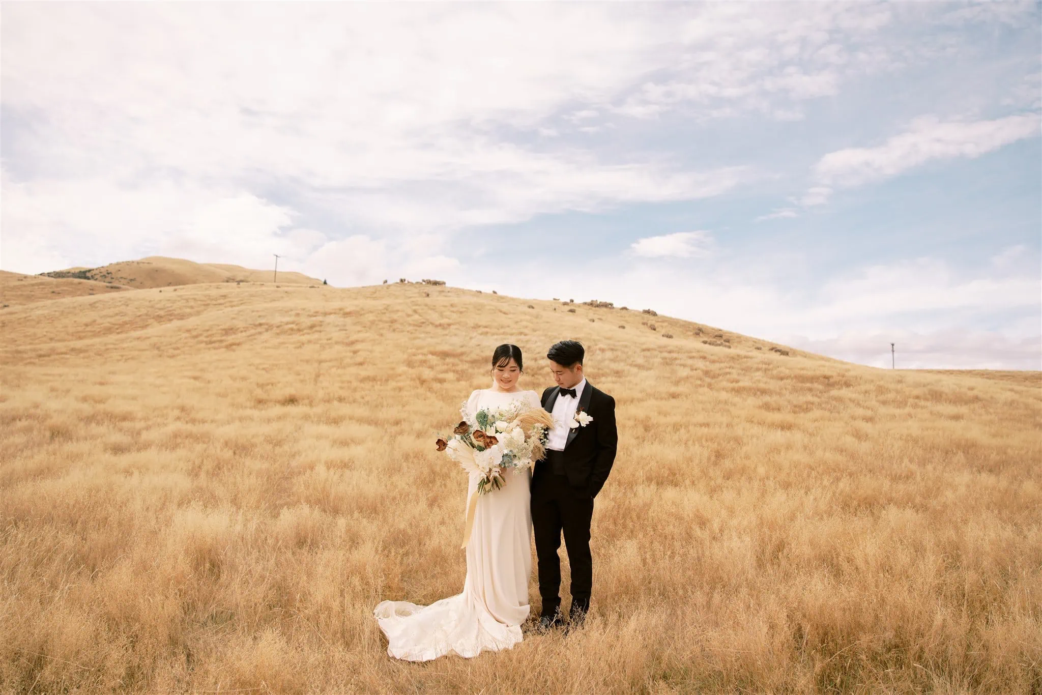 queenstown new zealand heli wedding elopement photographer videographer | A couple in formal attire stands together in a dry, grassy field under a partly cloudy sky, captured beautifully by a Queenstown wedding photographer. The woman holds a bouquet of flowers.