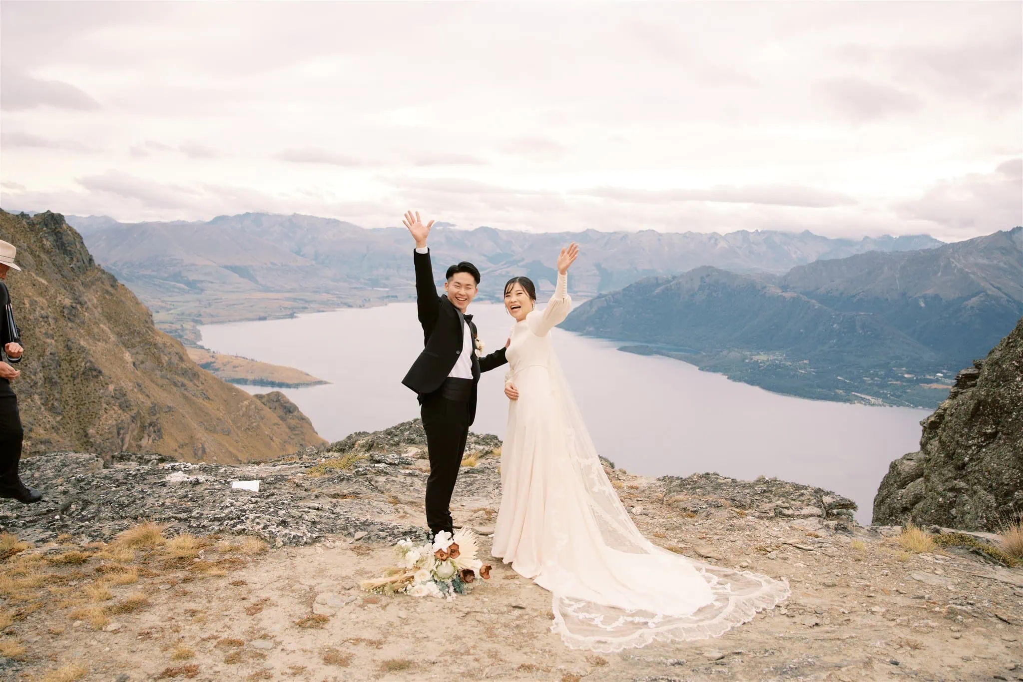 queenstown new zealand heli wedding elopement photographer videographer | A couple in wedding attire stands on a rocky overlook, waving at the camera with a lake and mountains in the background. Flowers are on the ground beside them. An additional person is visible in the distance, capturing this stunning moment—perhaps a Queenstown wedding photographer making magic happen.