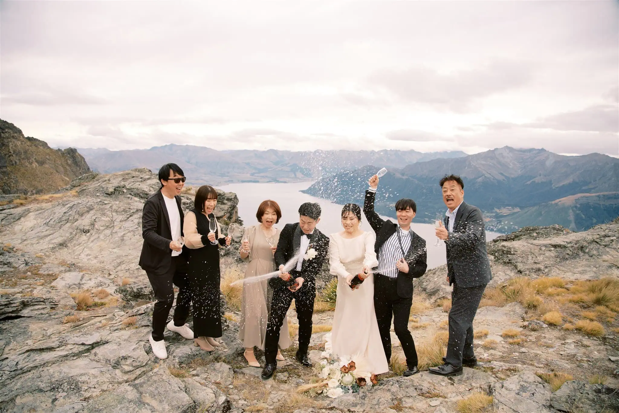 queenstown new zealand heli wedding elopement photographer videographer | A group of seven people, dressed in formal attire, celebrate outdoors on a rocky landscape with mountains in the background. They are opening champagne bottles, and some are cheering and posing for the photo, perfectly captured by a skilled Queenstown wedding photographer.