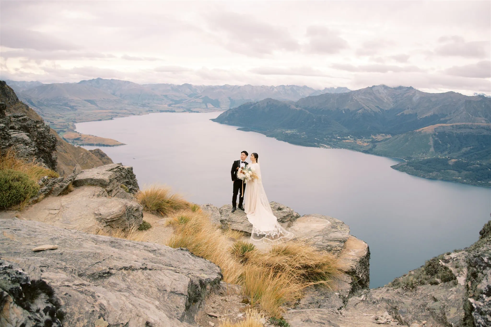 queenstown new zealand heli wedding elopement photographer videographer | A bride and groom stand on a rocky outcrop overlooking a lake and mountains, with the bride in a white dress and the groom in a black suit, holding a bouquet. Captured by a Queenstown wedding photographer, this moment is truly breathtaking.
