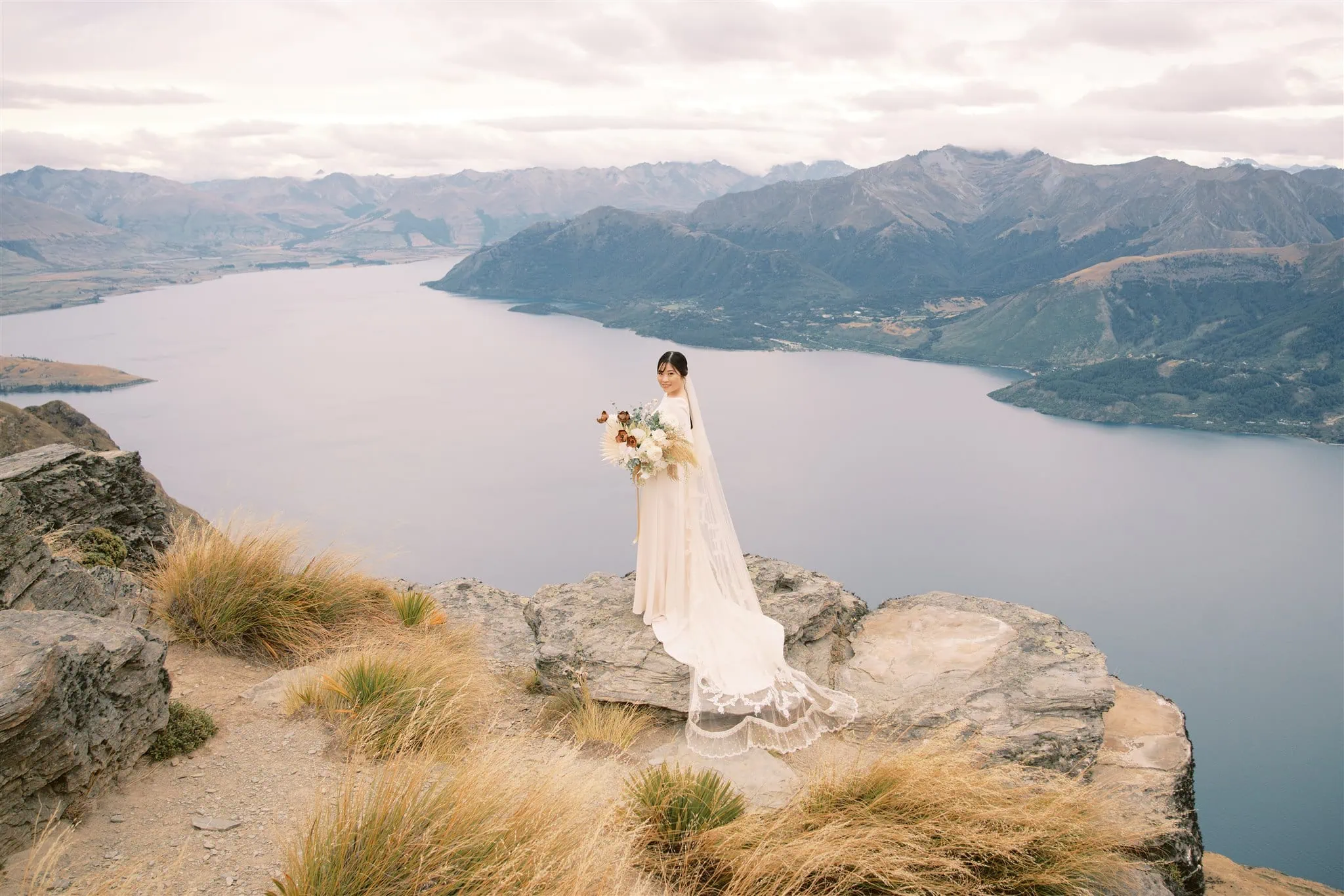 queenstown new zealand heli wedding elopement photographer videographer | A bride in a white dress and veil stands on a rocky cliff overlooking a large lake and mountain range, holding a bouquet of flowers. Captured by a Queenstown wedding photographer, this breathtaking moment combines natural beauty with timeless romance.