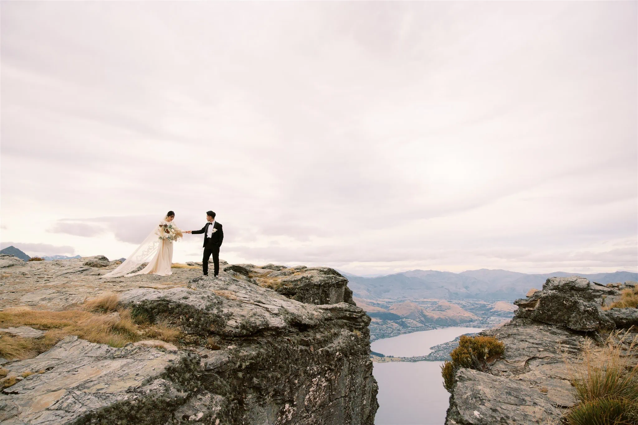 queenstown new zealand heli wedding elopement photographer videographer | A couple in wedding attire stands on a rocky cliff, captured beautifully by a Queenstown wedding photographer, overlooking a lake and mountains under a cloudy sky.
