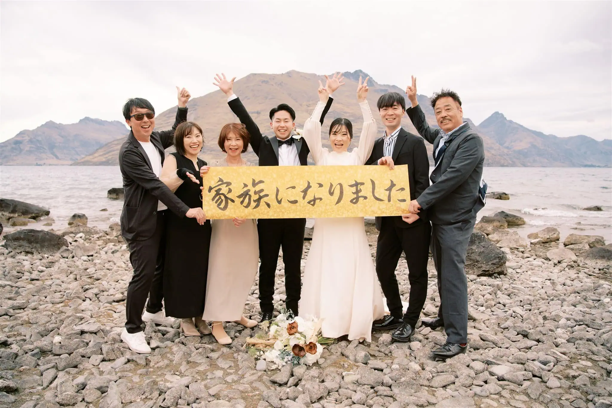 queenstown new zealand heli wedding elopement photographer videographer | A group of people standing on a rocky shoreline, likely captured by a Queenstown wedding photographer, hold a sign with Japanese text, against a stunning backdrop of mountains and water.