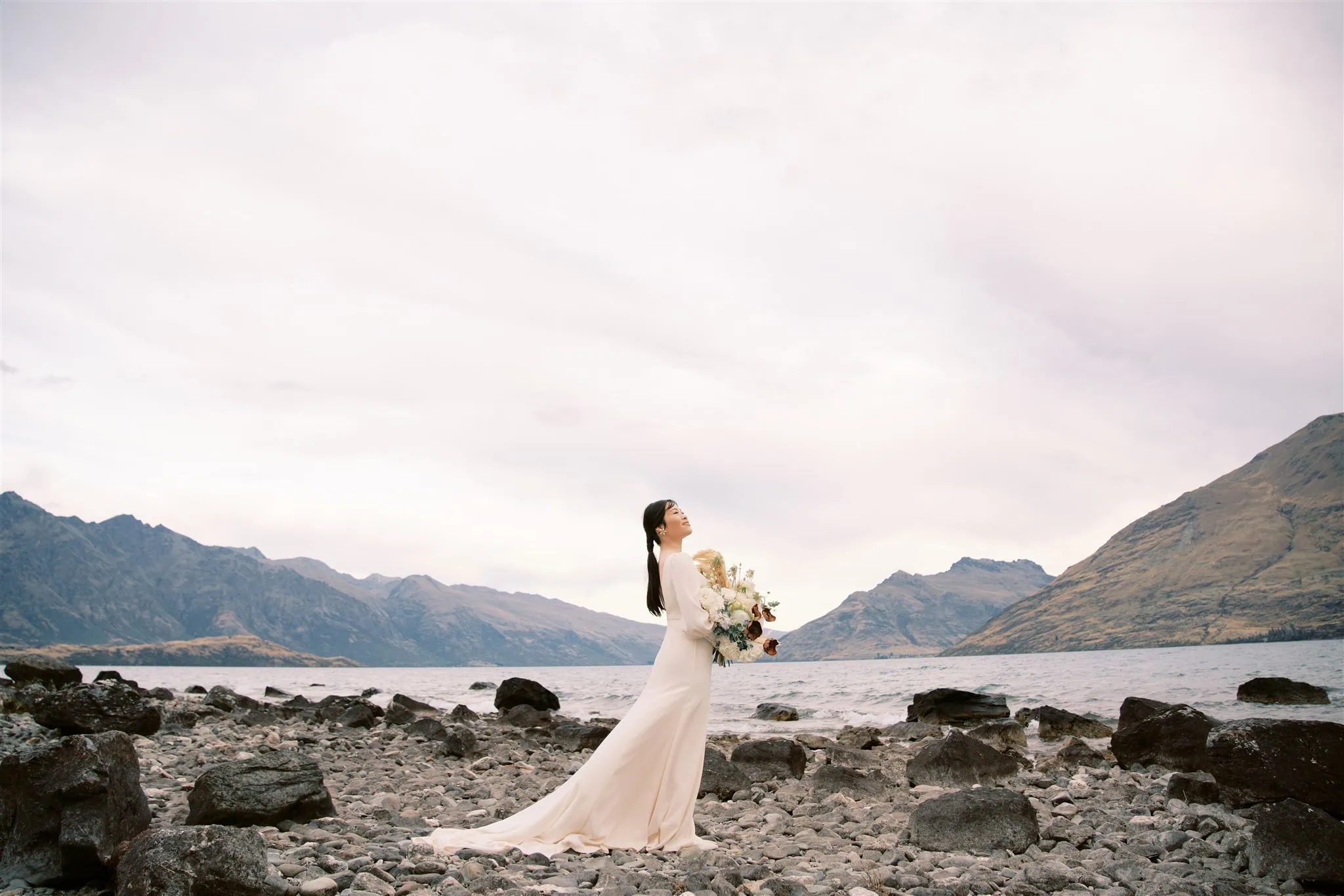 queenstown new zealand heli wedding elopement photographer videographer | A bride in a white dress stands on a rocky shoreline, holding a bouquet, with a backdrop of mountains and a cloudy sky—a moment perfectly captured by an expert Queenstown wedding photographer.