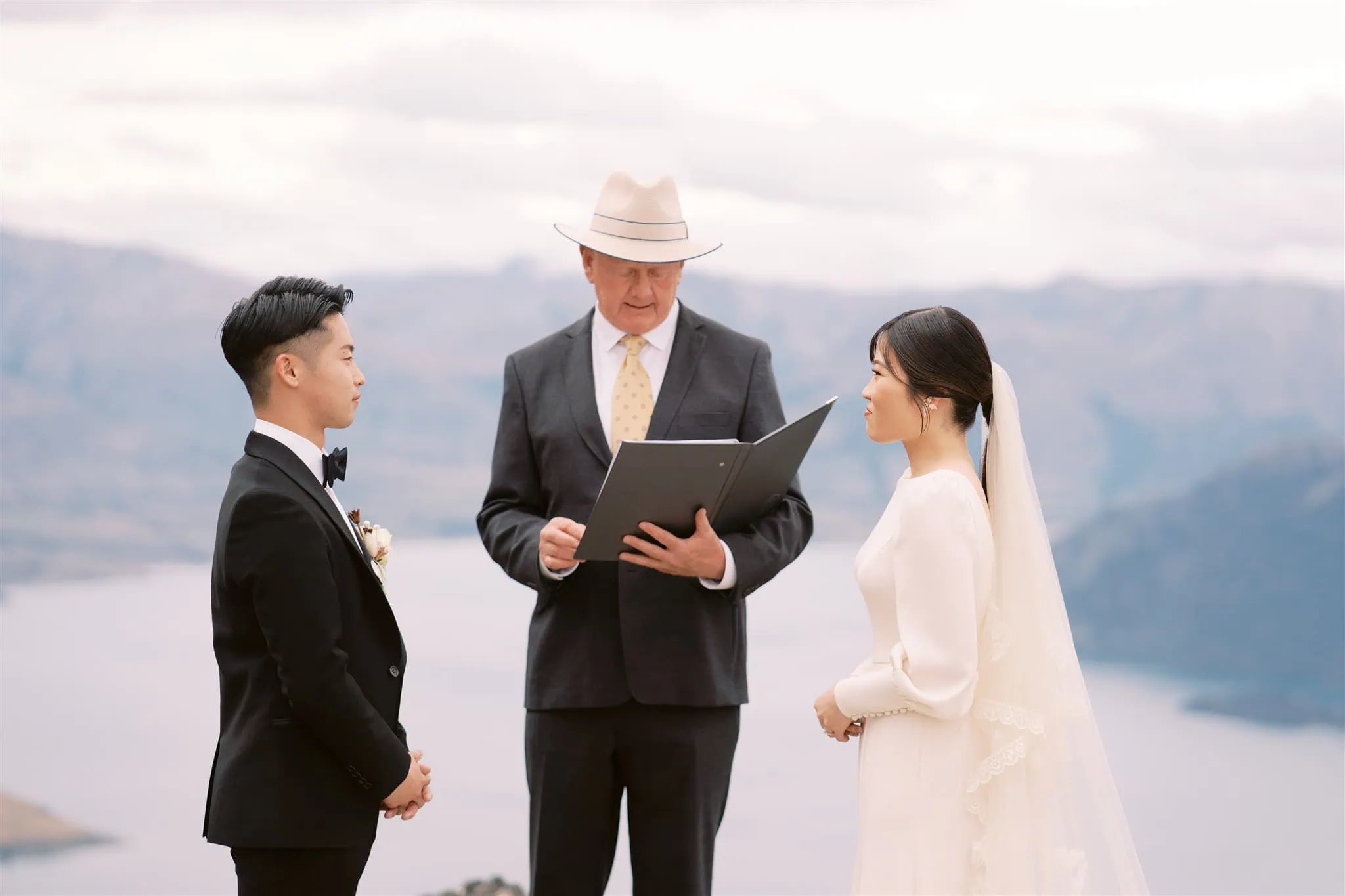 queenstown new zealand heli wedding elopement photographer videographer | A bride and groom stand facing each other during an outdoor ceremony officiated by a man in a suit and hat, with a scenic landscape of mountains and water in the background, beautifully captured by a Queenstown wedding photographer.