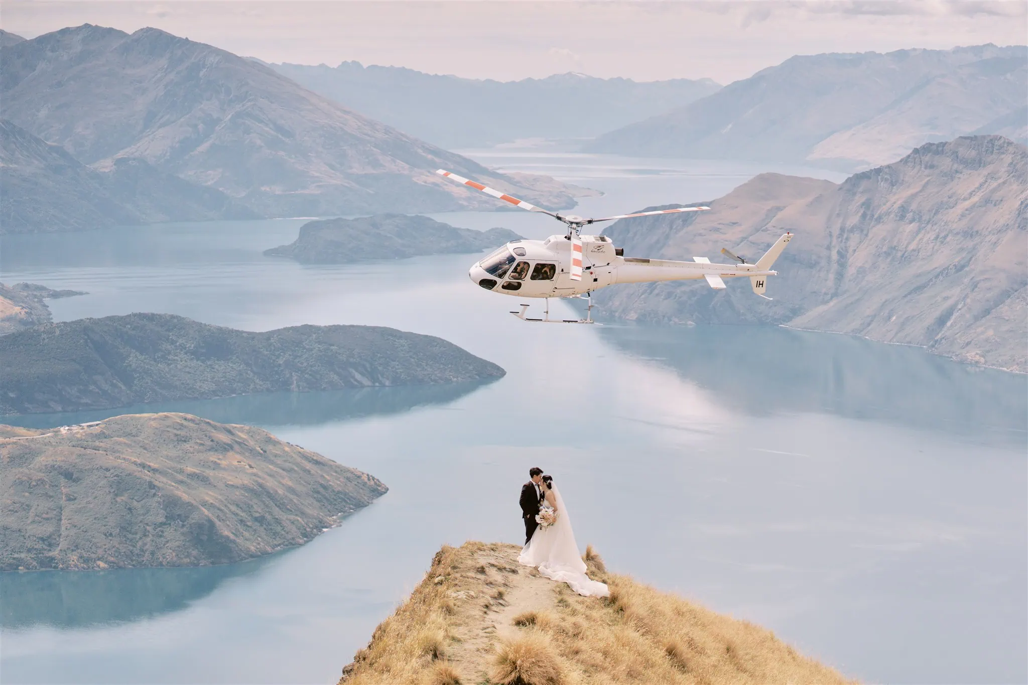 queenstown new zealand heli wedding elopement photographer videographer | A newlywed couple, Ryo & Tamano, standing on a hilltop with a helicopter flying nearby, overlooking a vast lake and mountainous landscape.