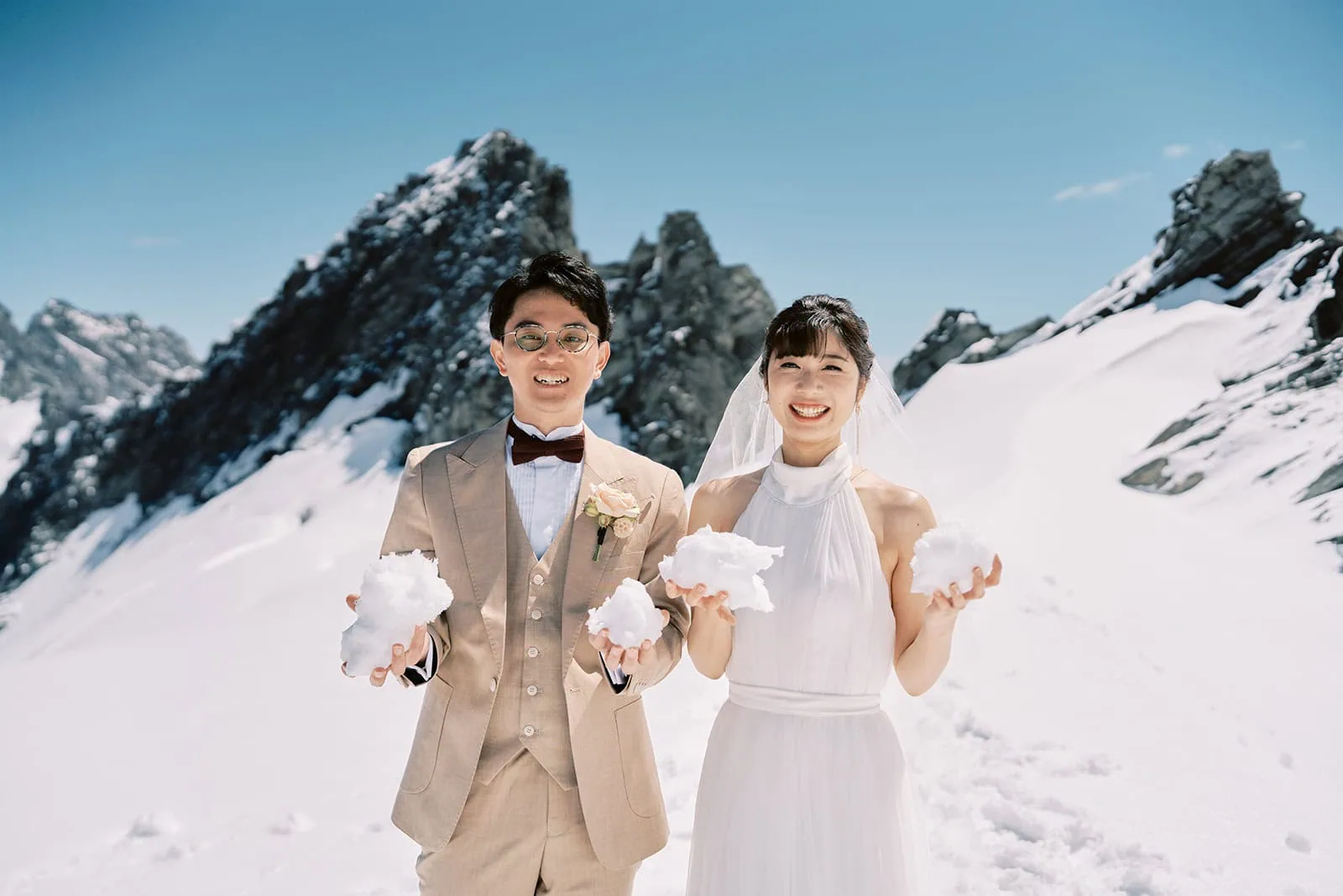 queenstown new zealand heli wedding elopement photographer videographer | A newlywed couple in wedding attire joyfully holding snowballs, standing against a backdrop of snow-covered mountains under a clear blue sky during their Queenstown Pre-Wedding Shoot.