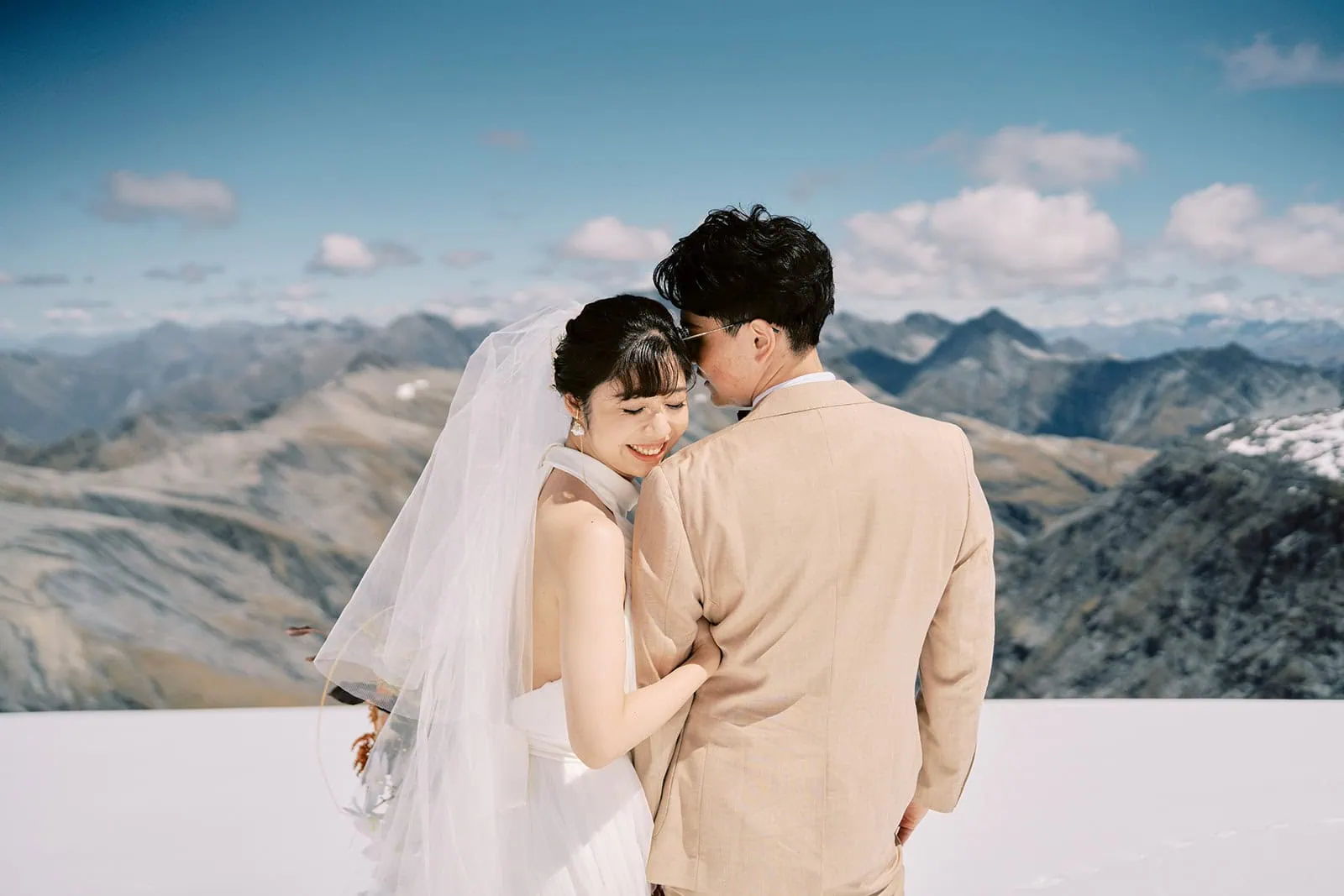 queenstown new zealand heli wedding elopement photographer videographer | A bride and groom embrace in a snowy mountain landscape during their Queenstown Pre-Wedding Shoot, with the bride smiling and the groom facing away from the camera.