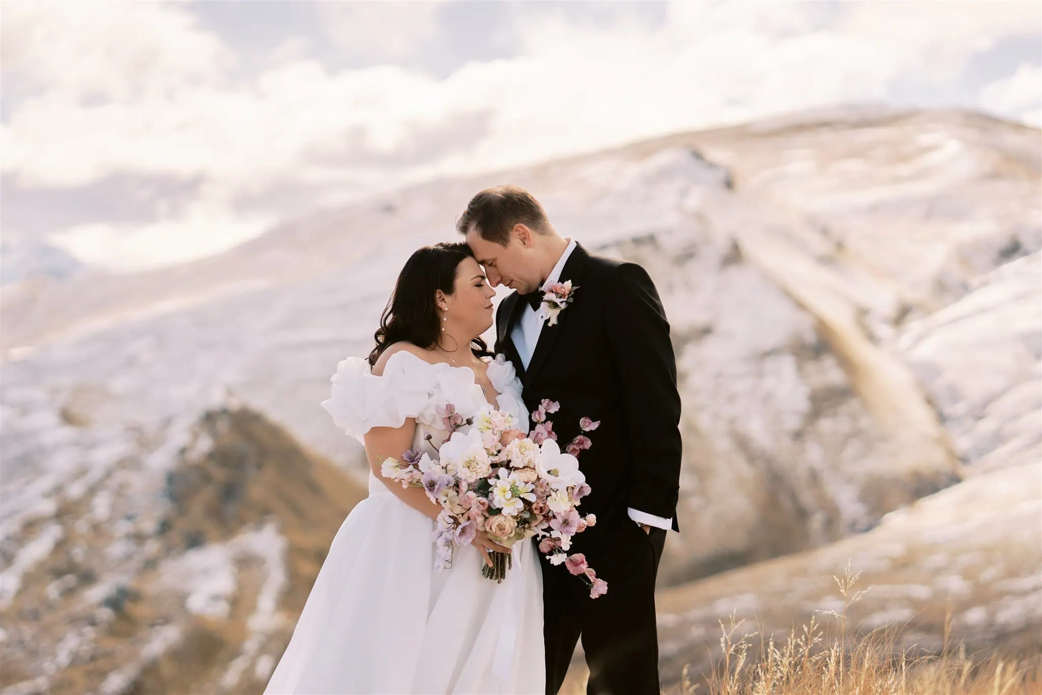 queenstown new zealand heli wedding elopement photographer videographer | Damien & Nanci stand close together amidst a mountainous landscape, the bride holding a bouquet of flowers. Snow-covered hills are visible in the background under a partly cloudy sky, captured beautifully by Queenstown Wedding Photography.