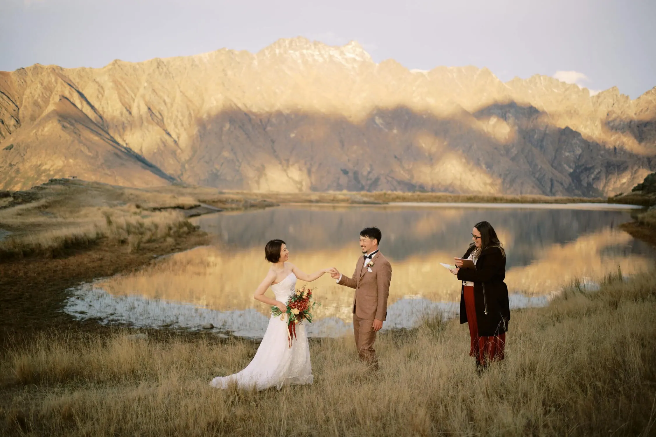 queenstown new zealand heli wedding elopement photographer videographer | A couple holds hands by a mountain lake during their outdoor wedding ceremony officiated by a third person, with mountains and calm water reflecting the scenic view in the background—a moment made even more magical by their Queenstown heli-wedding package.