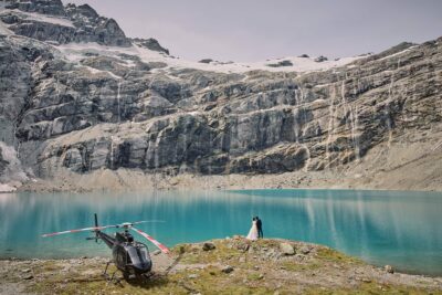 クイーンズタウン　テカポ　ニュージーランド　エロープメント 前撮り　フォトウェディング　結婚式 | クイーンズタウンのターコイズブルーの湖のそばにヘリコプターが停まっており、近くにはカップルが立っています。周囲は険しい岩の崖と滝に囲まれています。