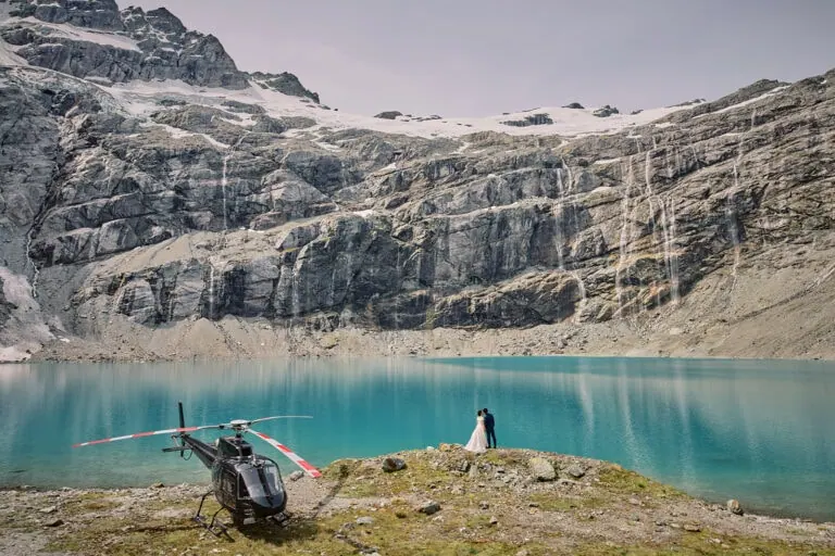 クイーンズタウン　テカポ　ニュージーランド　エロープメント 前撮り　フォトウェディング　結婚式 | クイーンズタウンのターコイズブルーの湖のそばにヘリコプターが停まっており、近くにはカップルが立っています。周囲は険しい岩の崖と滝に囲まれています。