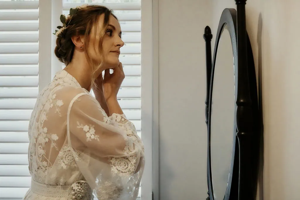 queenstown new zealand heli wedding elopement photographer videographer | A woman in a lace robe looks at her reflection in an oval mirror, her hand touching her face. White shutters cover the window behind her, capturing a serene, intimate moment perfect for elopement wedding photography in Queenstown.