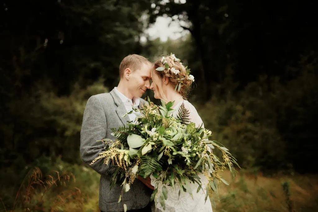 queenstown new zealand heli wedding elopement photographer videographer | A couple dressed in wedding attire stands close together, touching foreheads, with the bride holding a large bouquet of green and white flowers. The backdrop features lush greenery and trees, capturing the essence of Queenstown Elopement magic through stunning wedding photography.