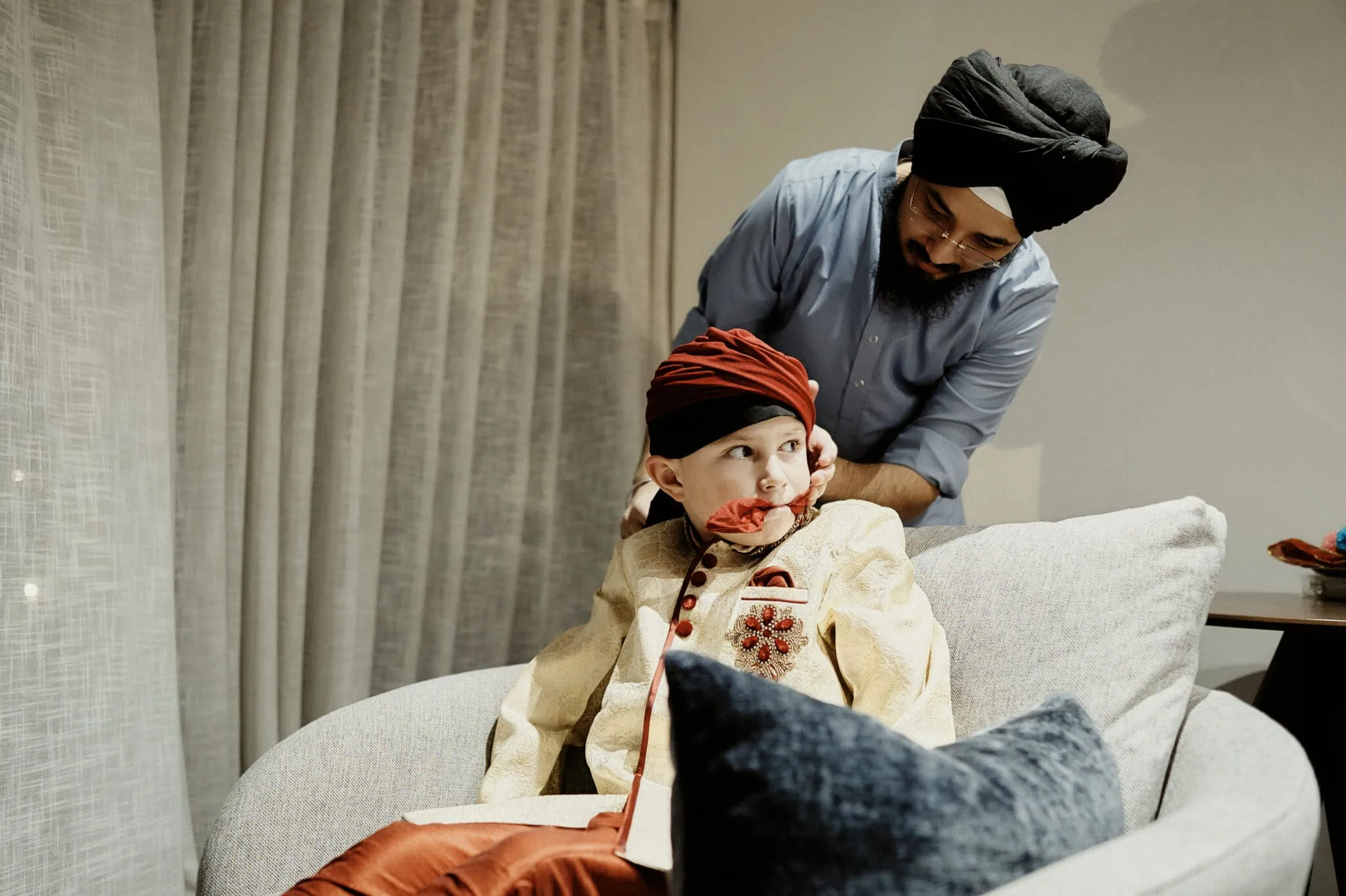 queenstown new zealand heli wedding elopement photographer videographer | A man helps a young boy wearing traditional attire and a turban sit comfortably on a grey chair during the Queenstown Elopement of Jasveen & Ben.