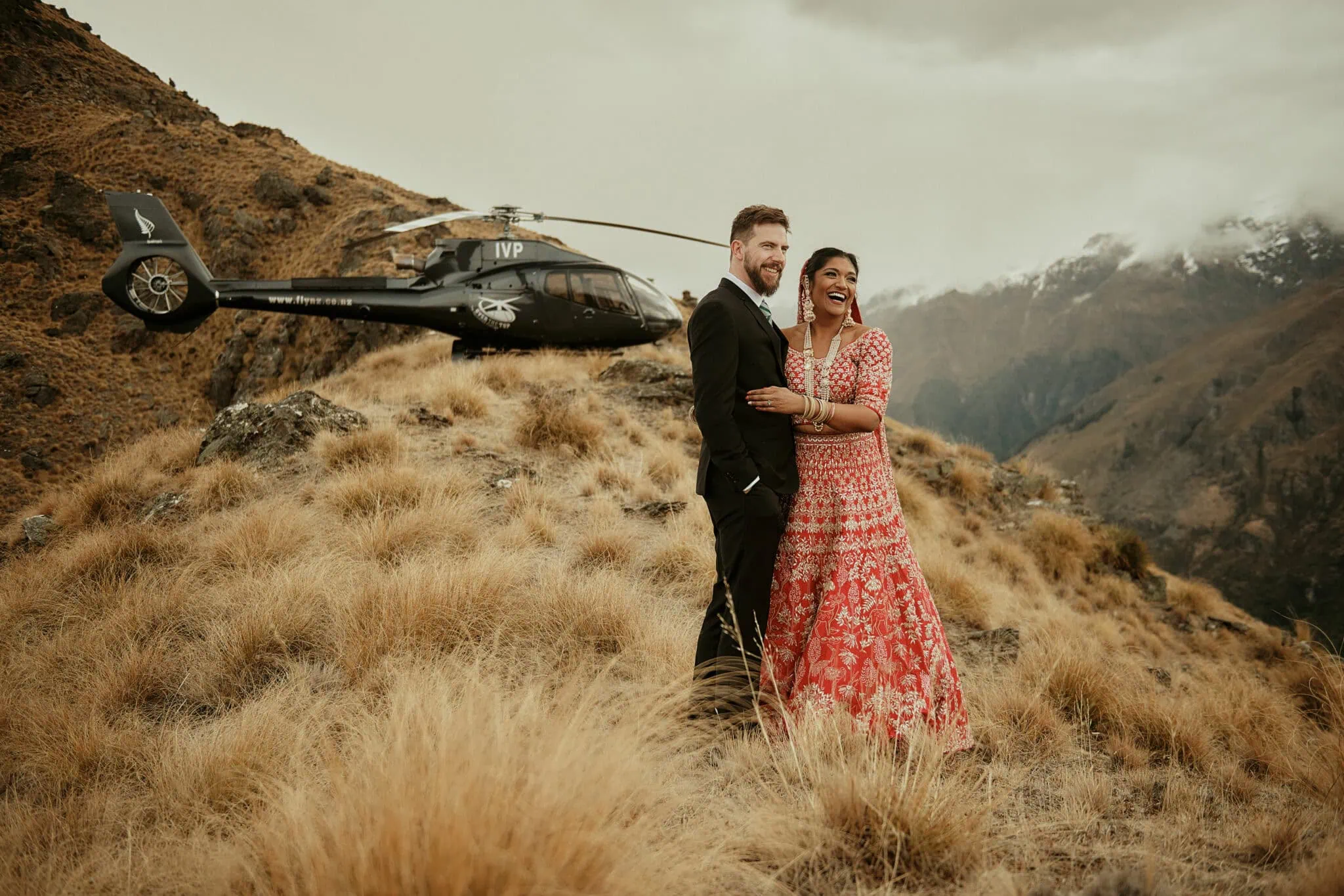 queenstown new zealand heli wedding elopement photographer videographer | Jasveen & Ben, elegantly dressed in formal wear, stand on a grassy hillside with a helicopter parked nearby and majestic mountains in the background. This stunning Queenstown elopement showcases incredible wedding photography amidst breathtaking scenery.