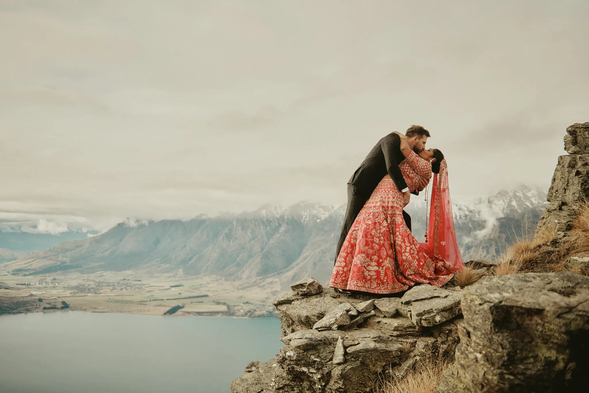 queenstown new zealand heli wedding elopement photographer videographer | A couple in formal attire shares a kiss at the edge of a rocky cliff with a scenic, mountainous landscape and a lake in the background, perfect for an elopement wedding photography session in Queenstown.