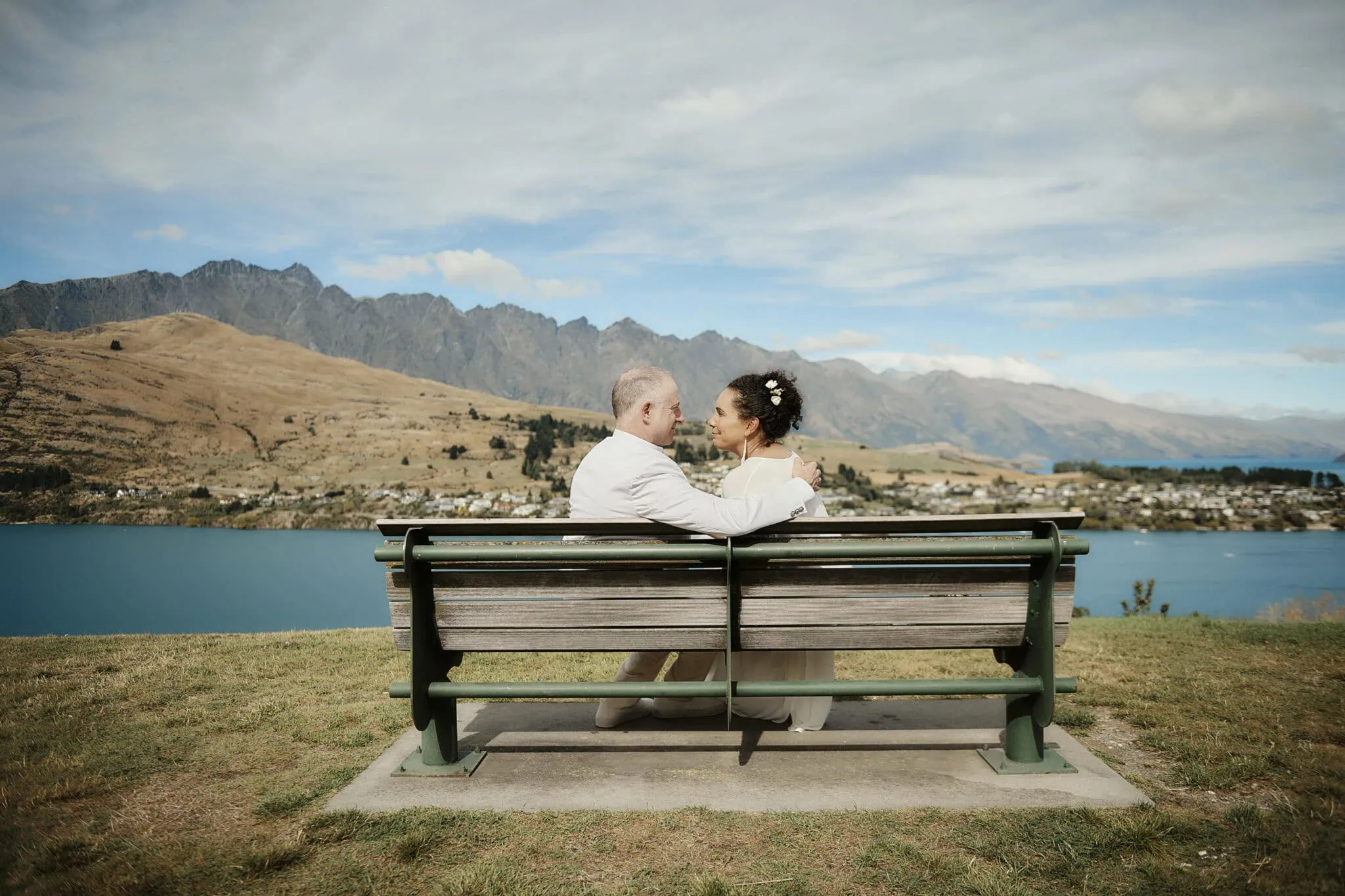 queenstown new zealand heli wedding elopement photographer videographer | A couple in wedding attire sits on a bench overlooking the scenic landscape of Queenstown with mountains, a lake, and a charming town in the background—a perfect setting for elopement wedding photography.