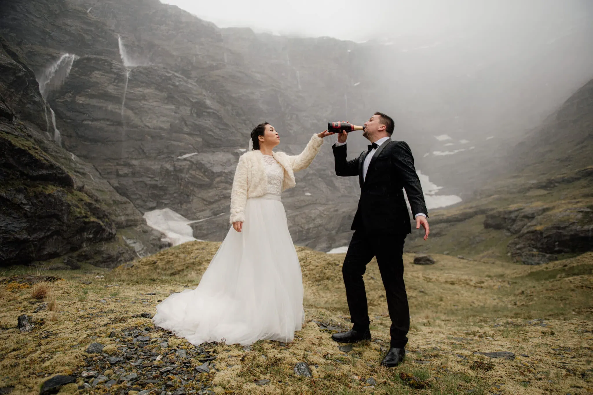 queenstown new zealand heli wedding elopement photographer videographer | Akiko & Dan, in wedding attire, stand on a foggy mountain in Queenstown; the bride holds a soda bottle while the groom drinks from it, capturing a whimsical moment in their pre-wedding photography.