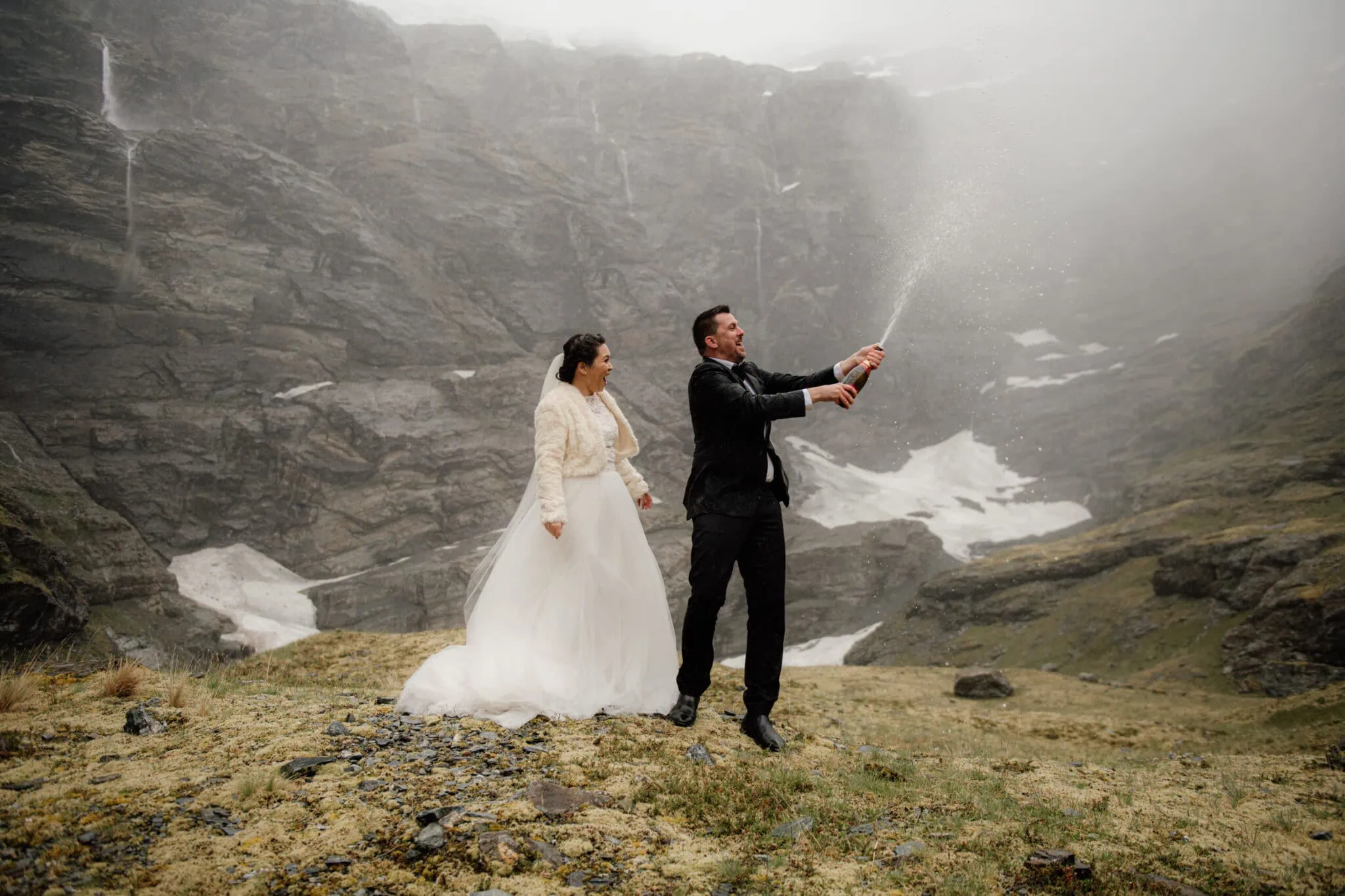 queenstown new zealand heli wedding elopement photographer videographer | A bride and groom in formal attire stand in a misty mountainous landscape, where the groom is popping a champagne bottle, and the bride watches. Snow patches and rugged cliffs are visible behind them in this stunning Queenstown Pre-Wedding Photography session with Akiko & Dan.