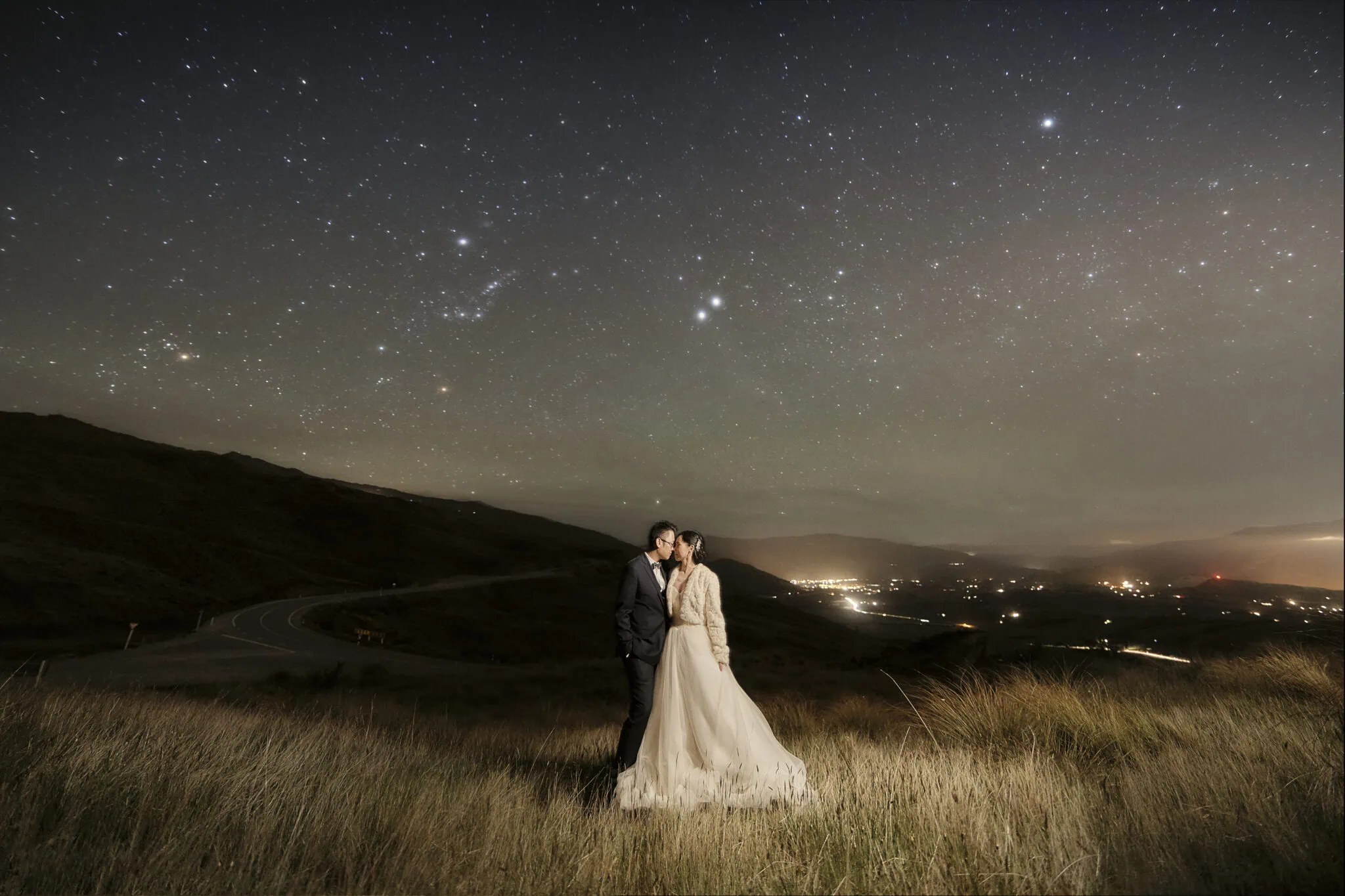 queenstown new zealand heli wedding elopement photographer videographer | A couple kissing under a starry night sky in a grassy, hilly area with city lights visible in the distance, captured perfectly in a Queenstown pre-wedding photography session.