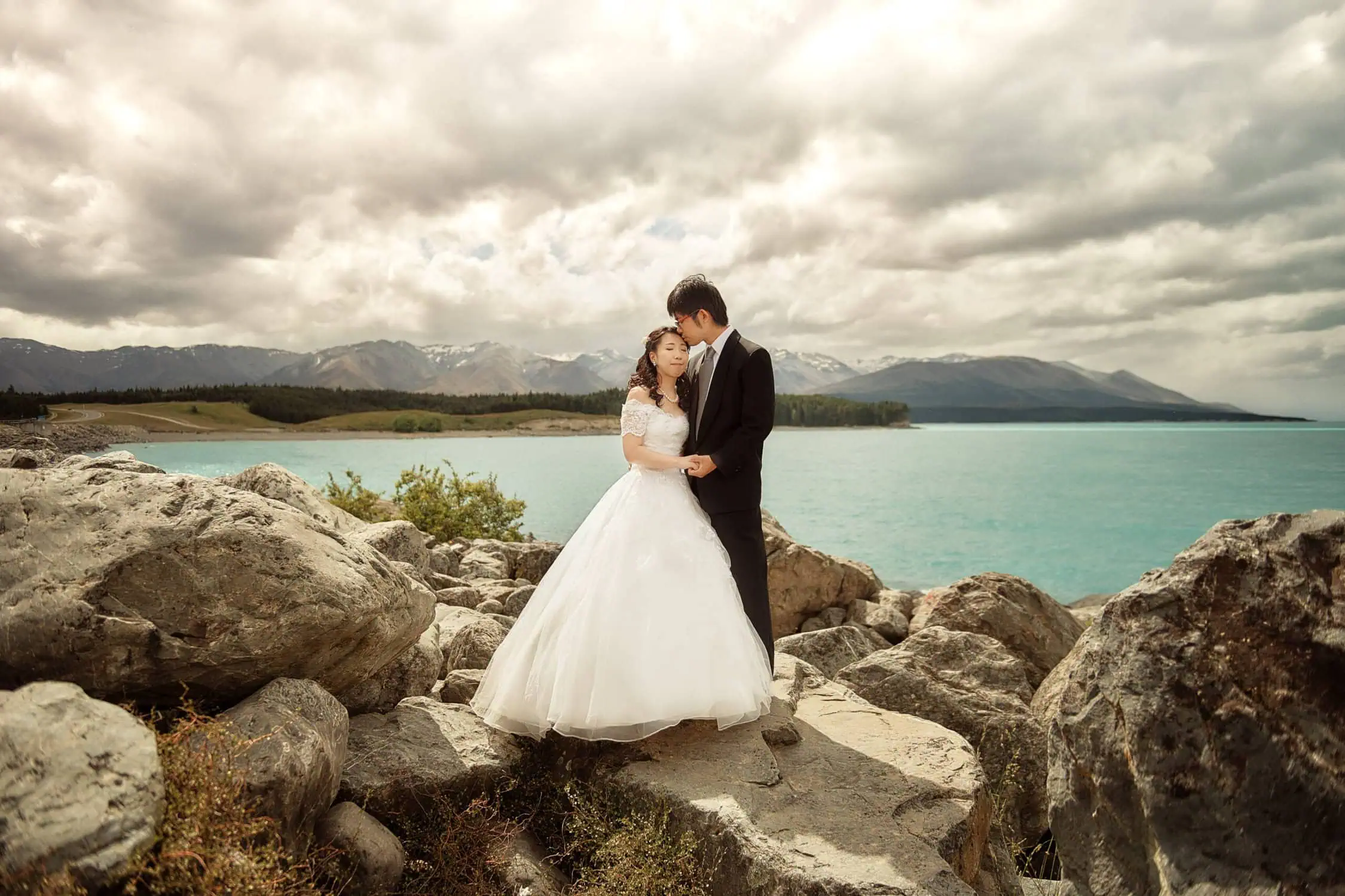 queenstown new zealand heli wedding elopement photographer videographer | A bride and groom, Eri & Youhei, stand on rocky terrain by a turquoise lake, with mountains and a cloudy sky in the background—a perfect Queenstown pre-wedding photography setting.