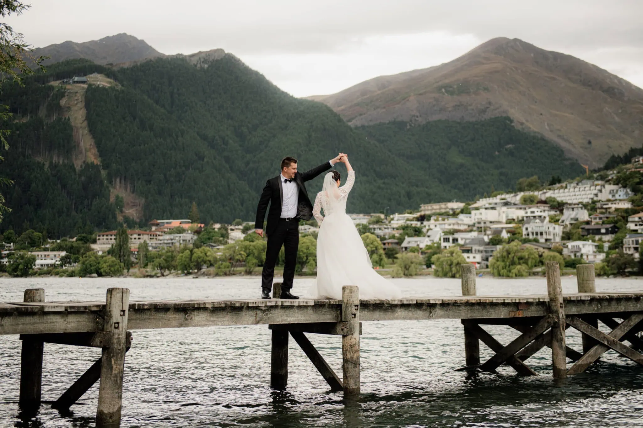 queenstown new zealand heli wedding elopement photographer videographer | Akiko & Dan, dressed in formal attire, dance gracefully on a wooden dock by a lake with mountains and houses in the background. This enchanting scene captures the elegance of Queenstown Pre-Wedding Photography.