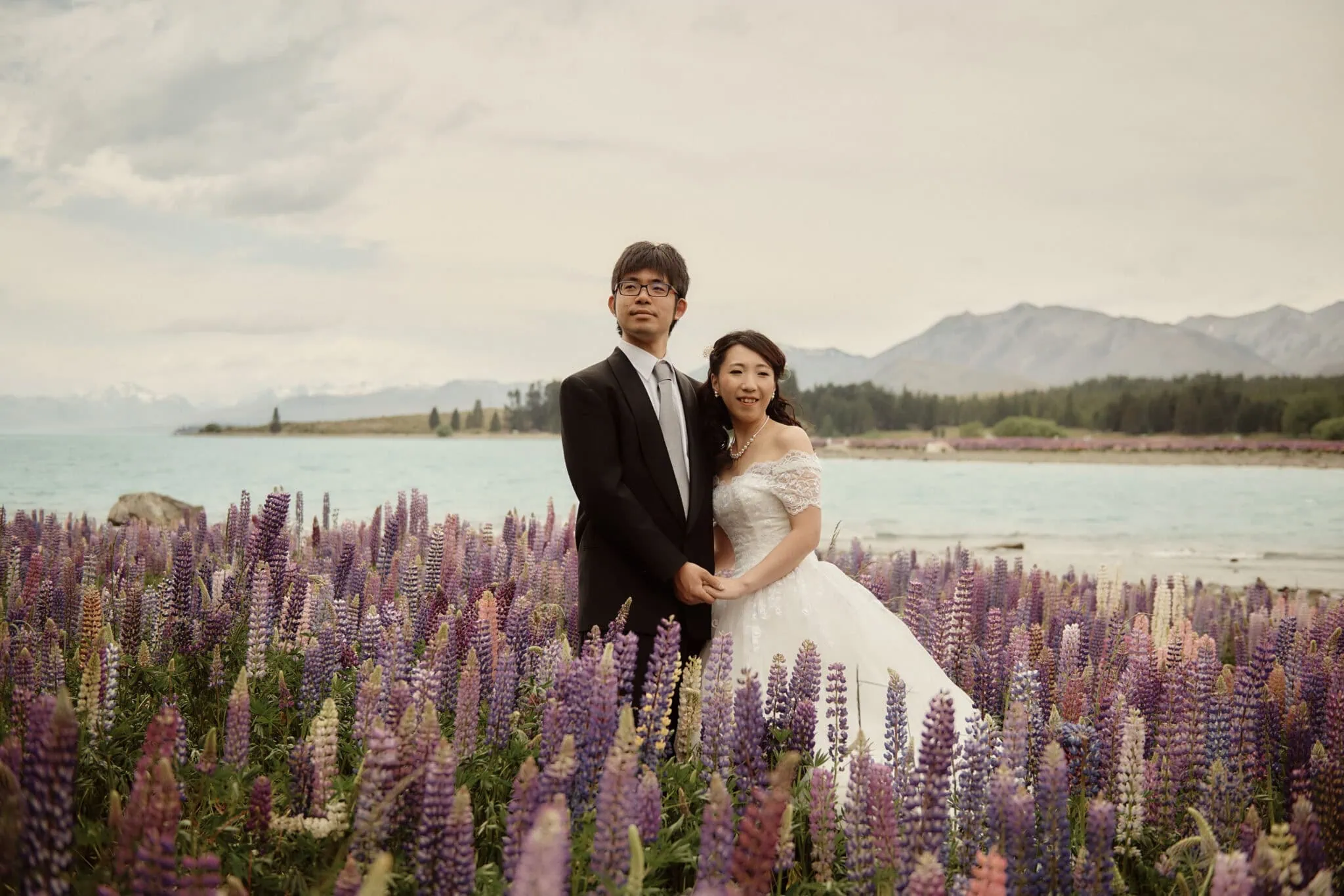 queenstown new zealand heli wedding elopement photographer videographer | A couple in wedding attire stands in a field of purple lupines with a lake and mountains in the background, capturing the essence of Queenstown Pre-wedding Photography.
