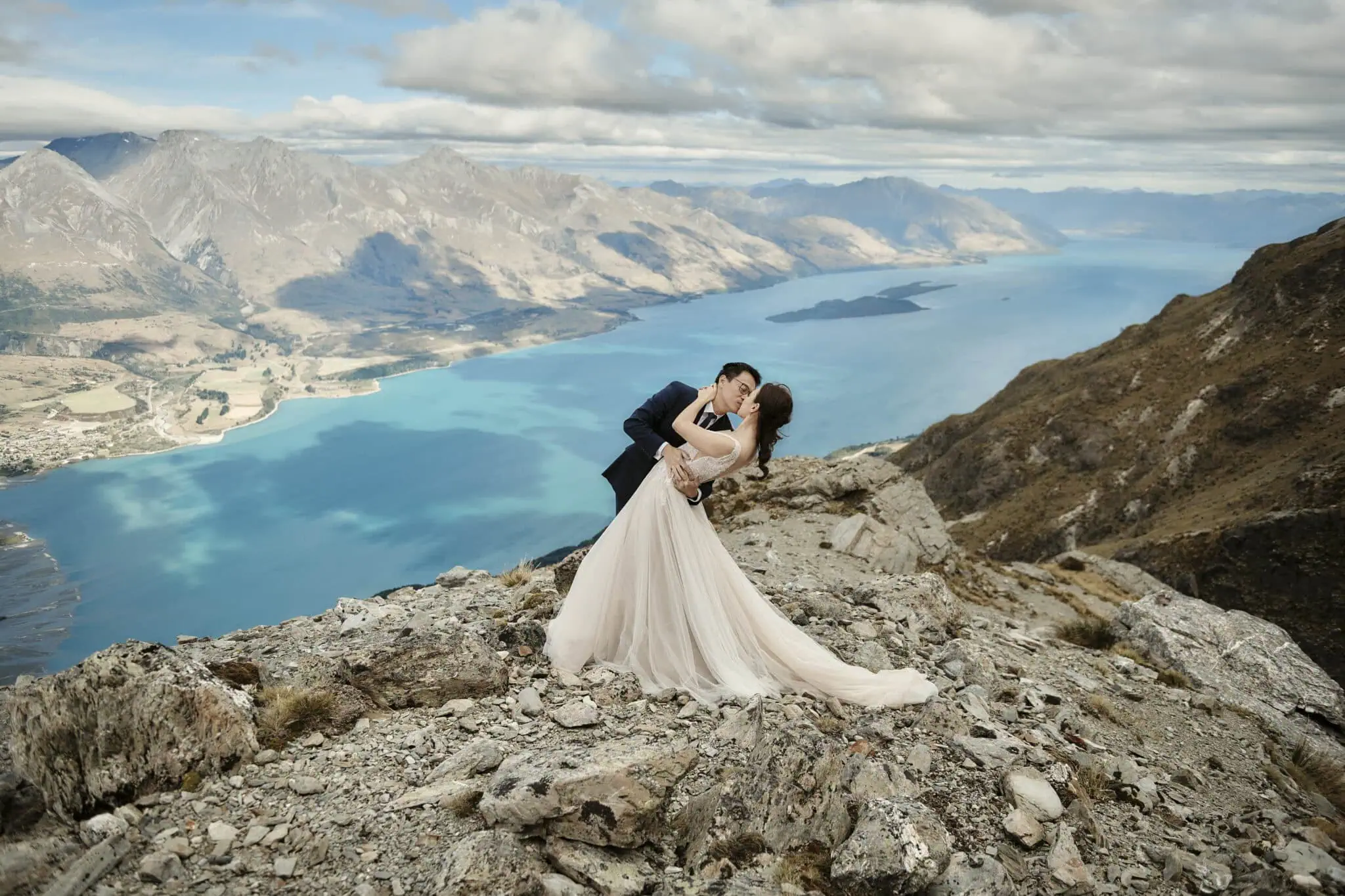queenstown new zealand heli wedding elopement photographer videographer | Mindy & Will, dressed in elegant wedding attire, share a tender dance on a rocky mountain cliff in Queenstown. Overlooking a scenic lake and majestic mountain range under a partially cloudy sky, their pre-wedding shoot captures the essence of romance and natural beauty.