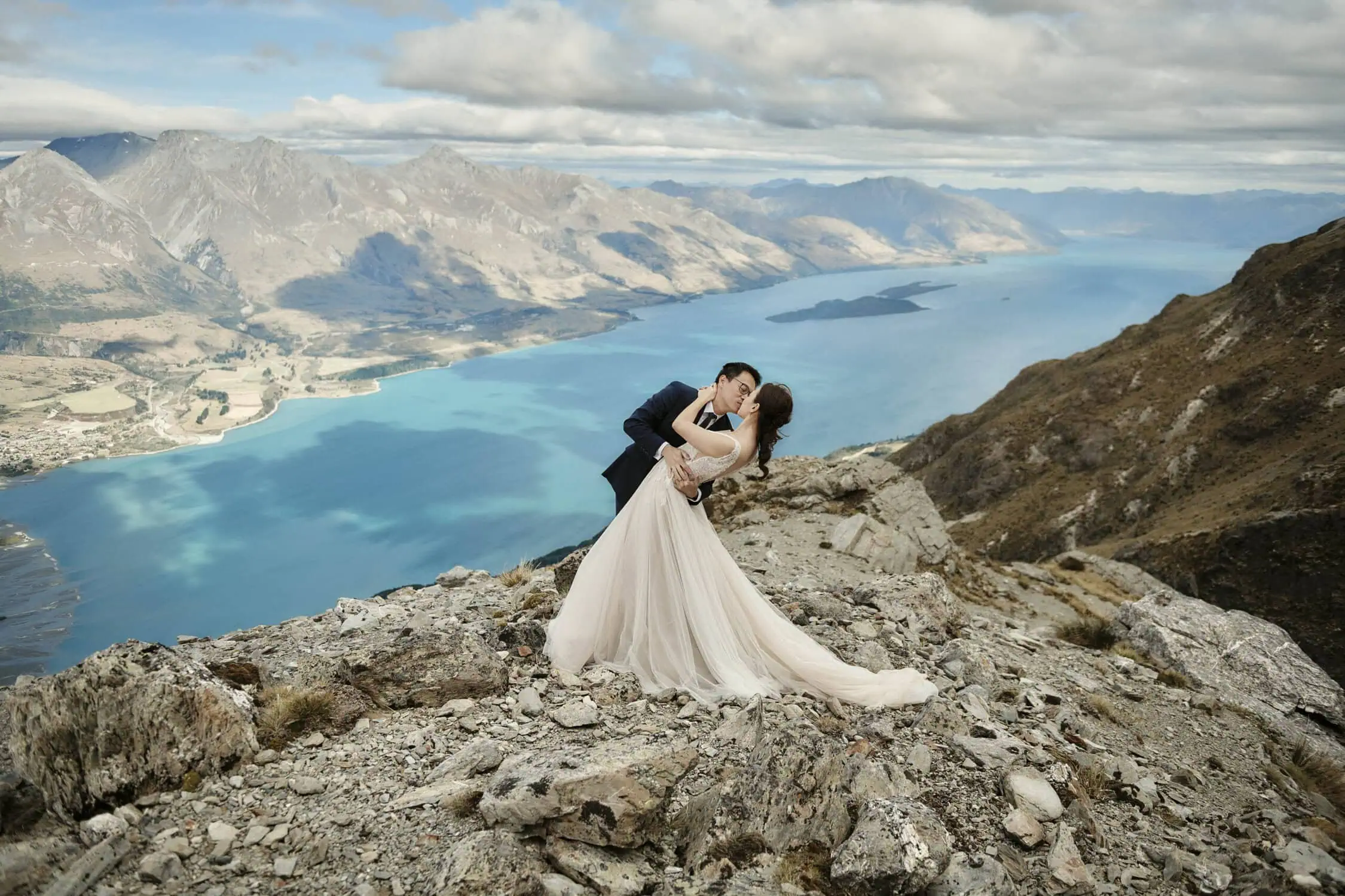 queenstown new zealand heli wedding elopement photographer videographer | Mindy & Will, dressed in elegant wedding attire, share a tender dance on a rocky mountain cliff in Queenstown. Overlooking a scenic lake and majestic mountain range under a partially cloudy sky, their pre-wedding shoot captures the essence of romance and natural beauty.