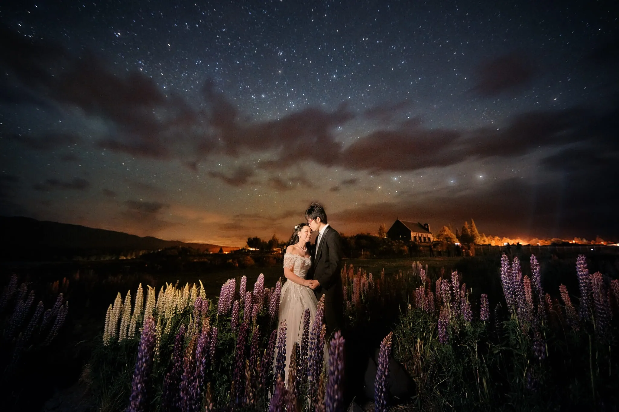 queenstown new zealand heli wedding elopement photographer videographer | A couple, Eri & Youhei, embraces in a field of lupine flowers under a starry night sky with scattered clouds and distant lights, capturing a magical moment in Queenstown for their pre-wedding photography.