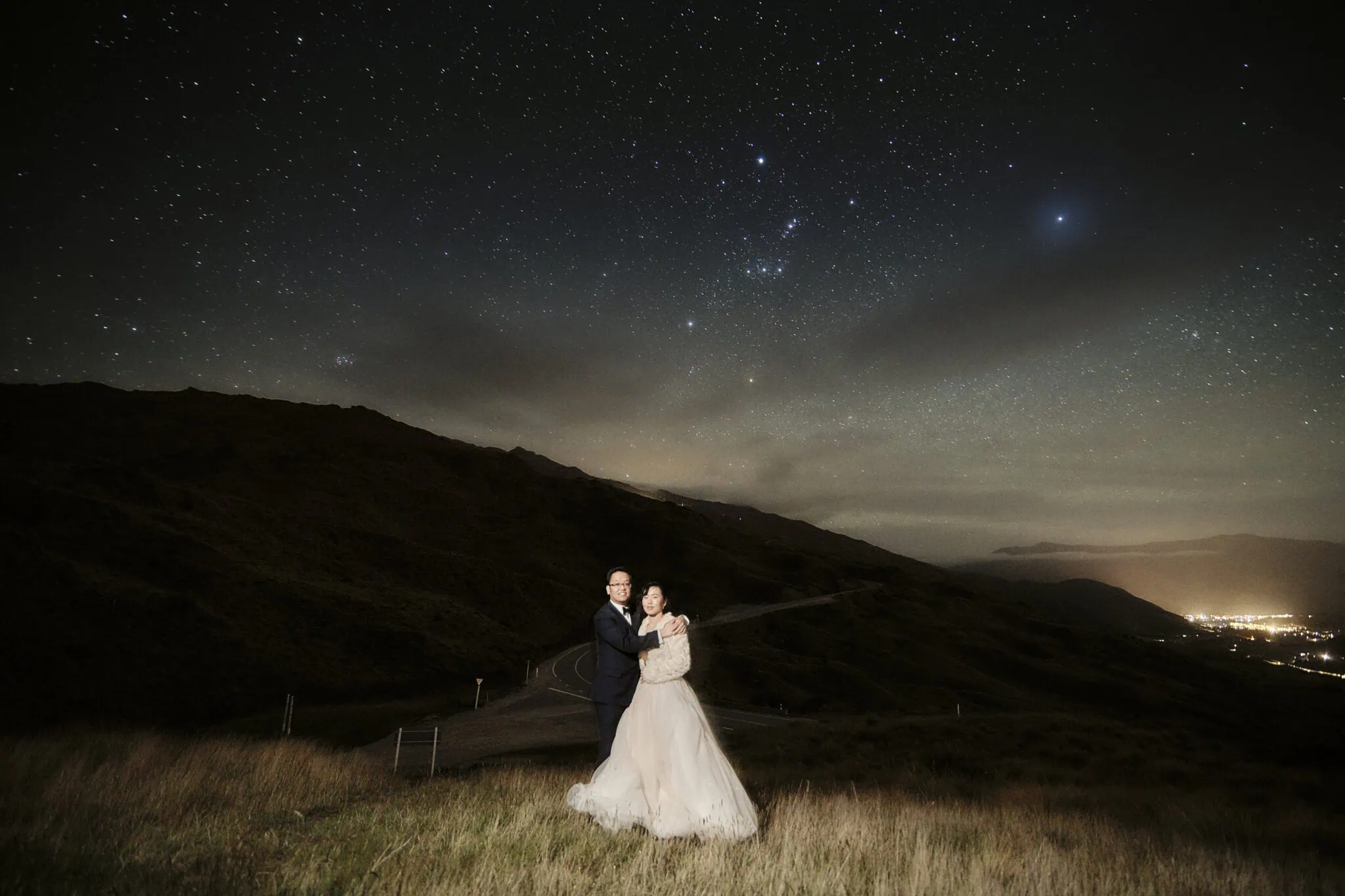 queenstown new zealand heli wedding elopement photographer videographer | A couple, Kristin & Shijun, dressed in wedding attire pose on a grassy hill in Queenstown under a star-filled night sky with distant city lights on the horizon.